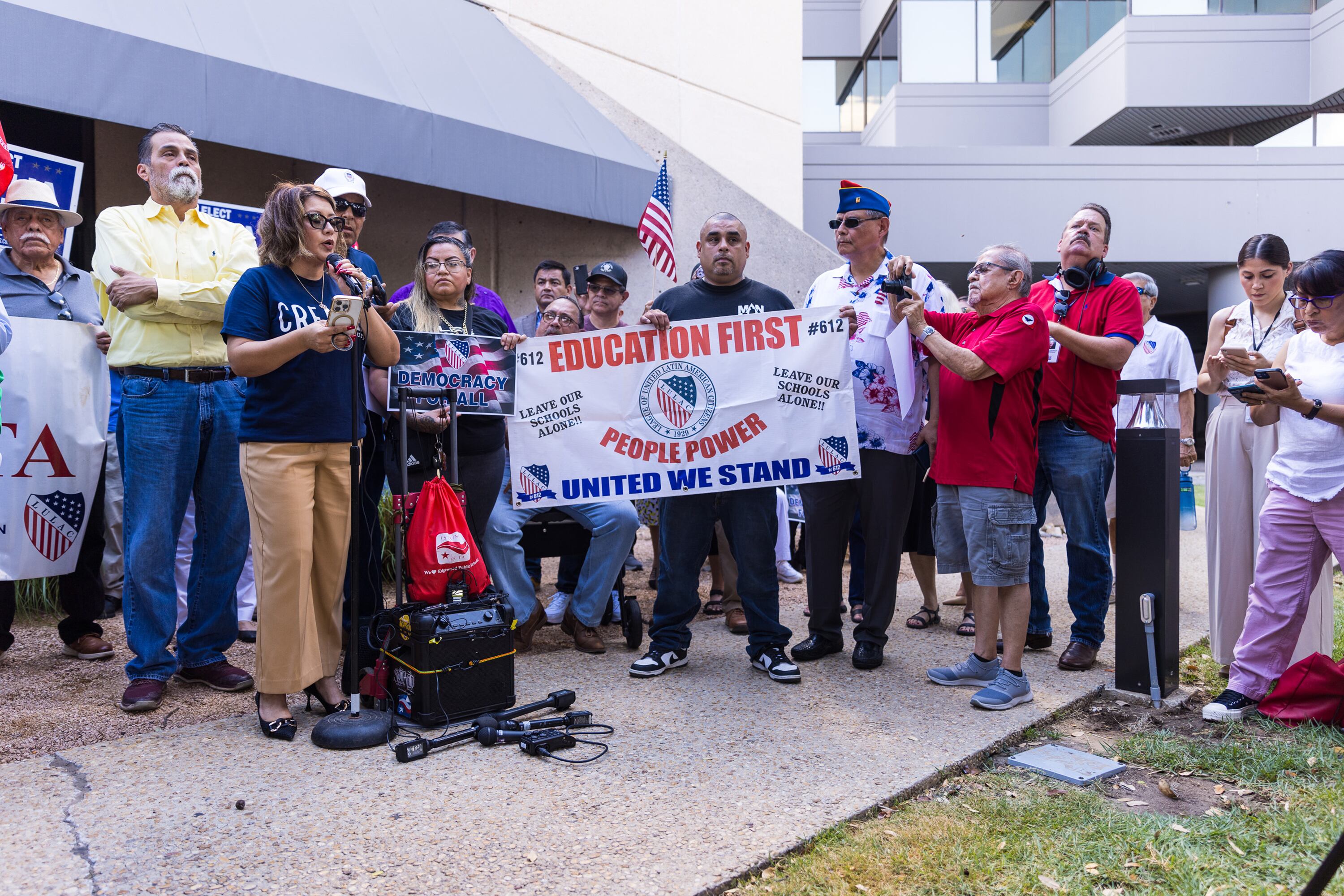 A group of people stand around a woman speaking into a microphone and portable speaker. One man holds a banner reading, "Education First. People Power. United We Stand."