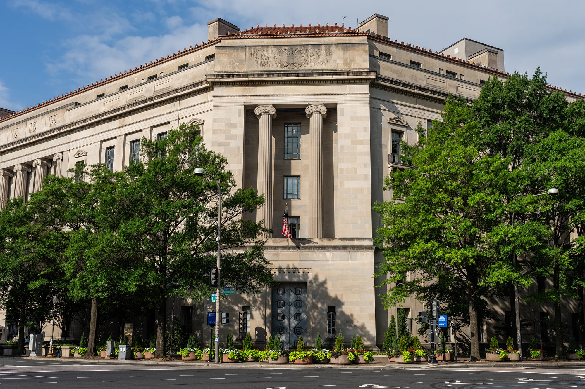A photograph of a large tan stone building with green trees out front and a person walking by.