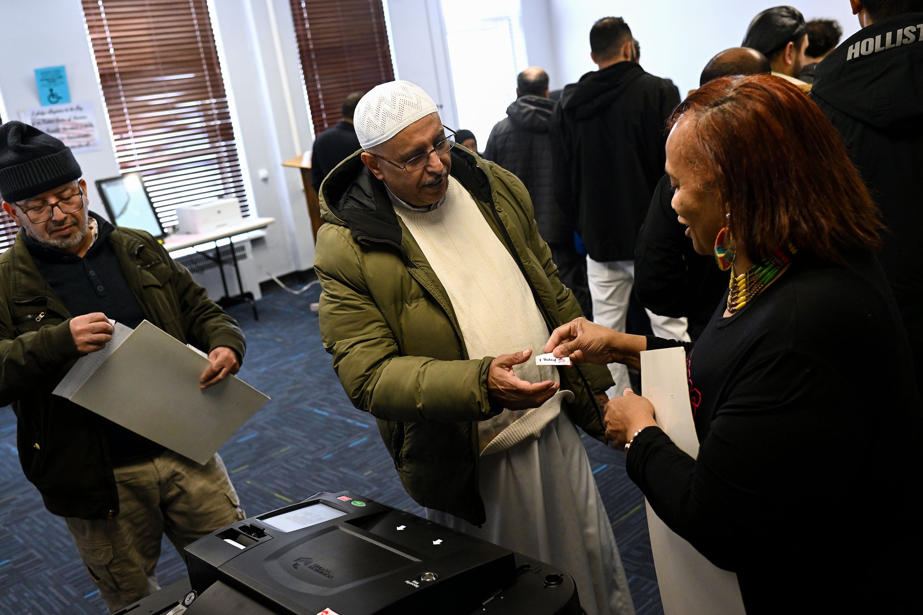 Three people are in view standing in a room with dark carpet. One person on the far right, hands an "I voted" sticker to the person in the middle. A couple of people are in the background.