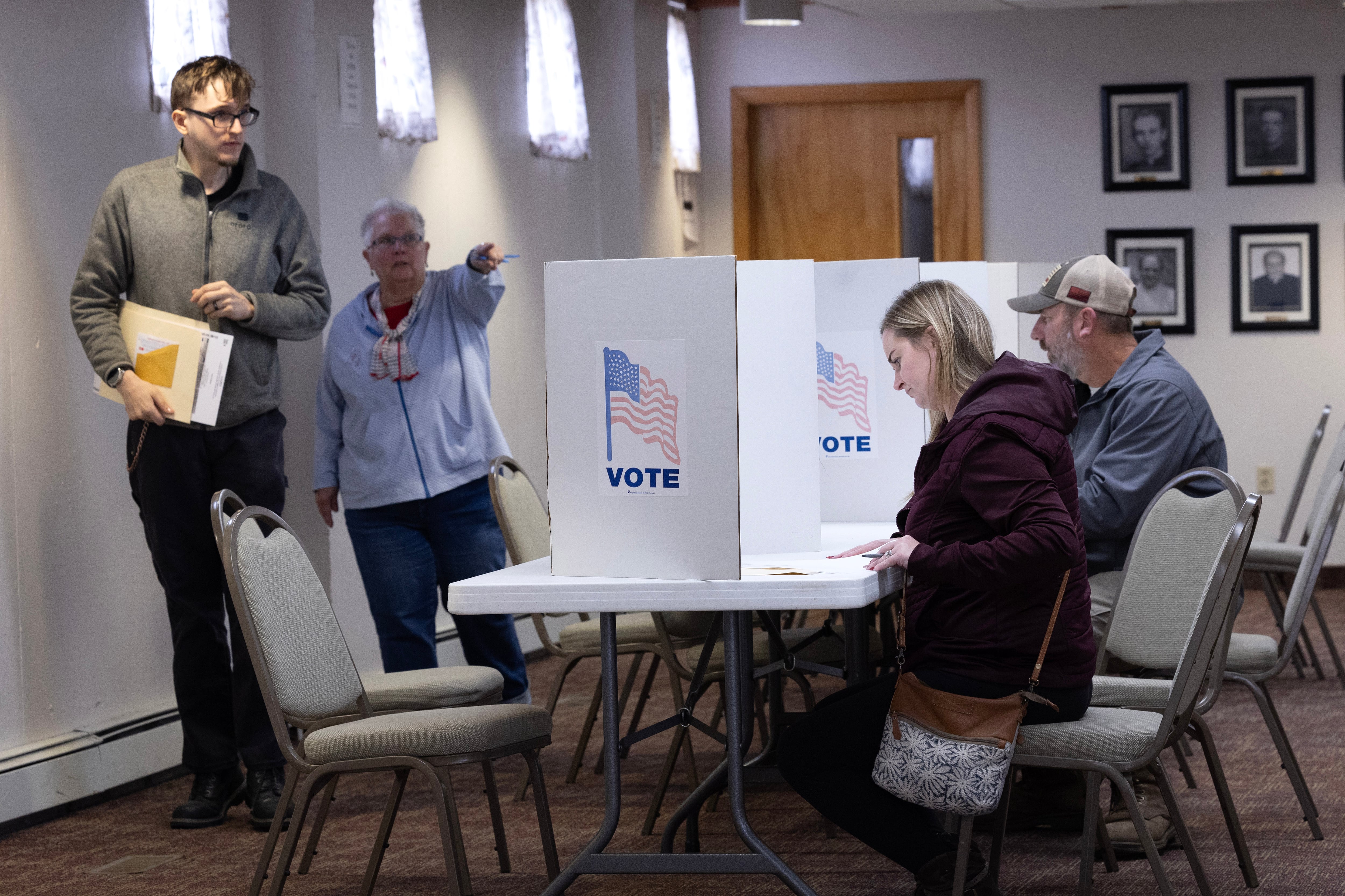 A woman in a light blue top points as a man in a light greenish jacket walks in front of her. Two people are at voting booths at right.