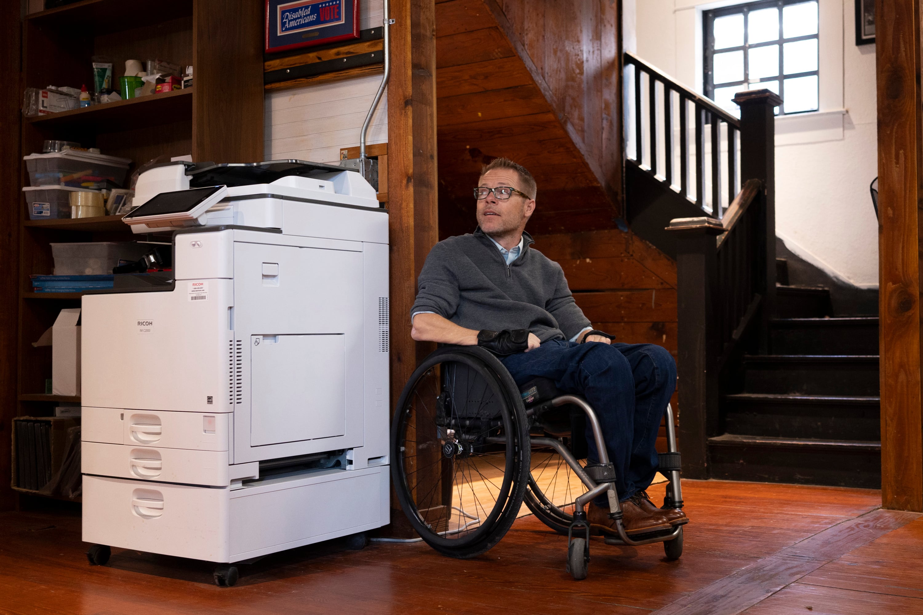 A man in a wheelchair poses for a photo inside an office near a printer