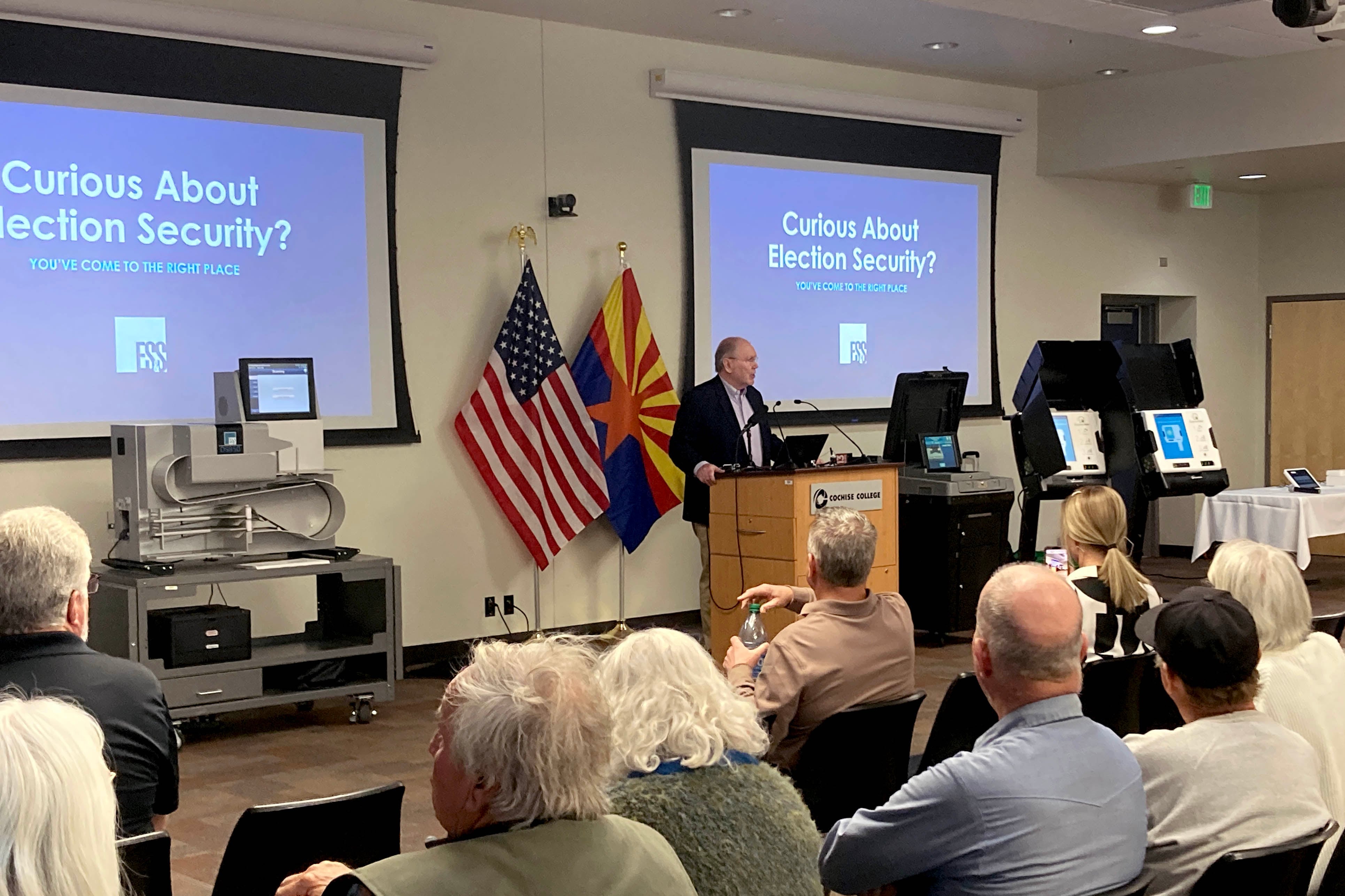 A man wearing a suit stands behind a podium and in front of several rows of people sitting in chairs. There are two projector screens hanging from the wall in the background and reads "Curious about election security."