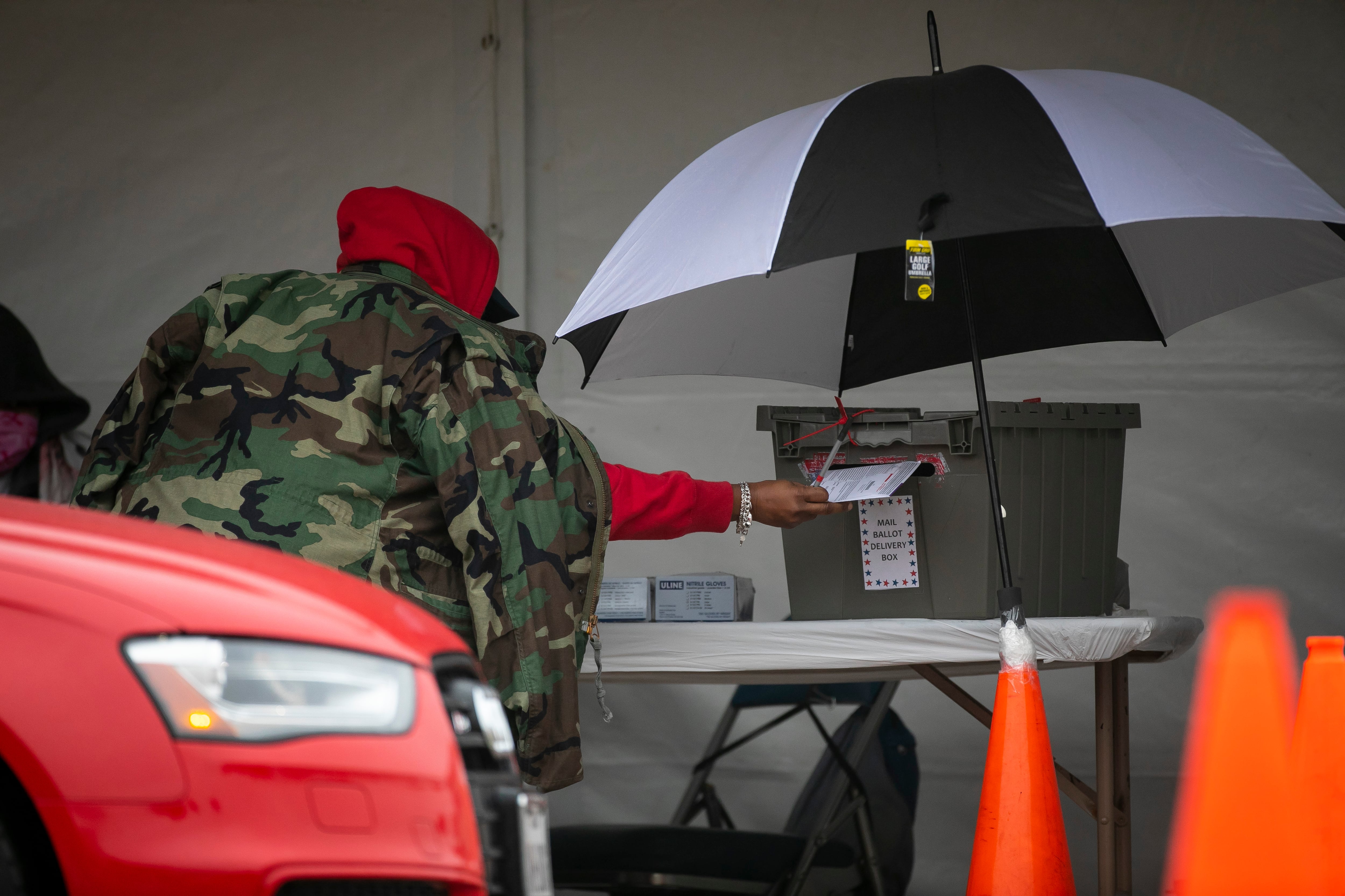 A photograph of a person wearing a red sweater covered by a camo jacket, reaches to drop off their ballot under an unbrella.