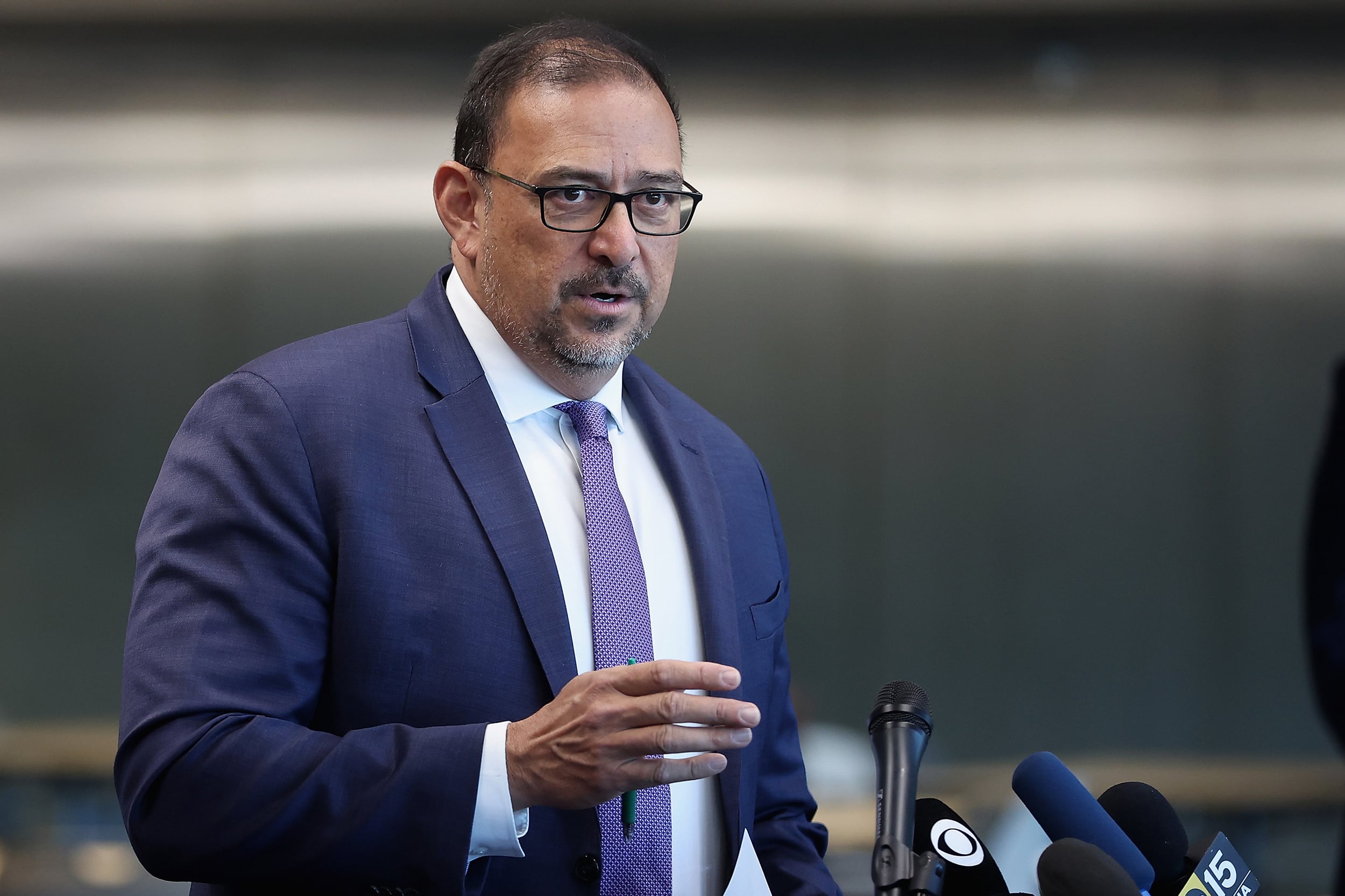 A photograph of a man in a suit with glasses speaking behind a podium with a few microphones.