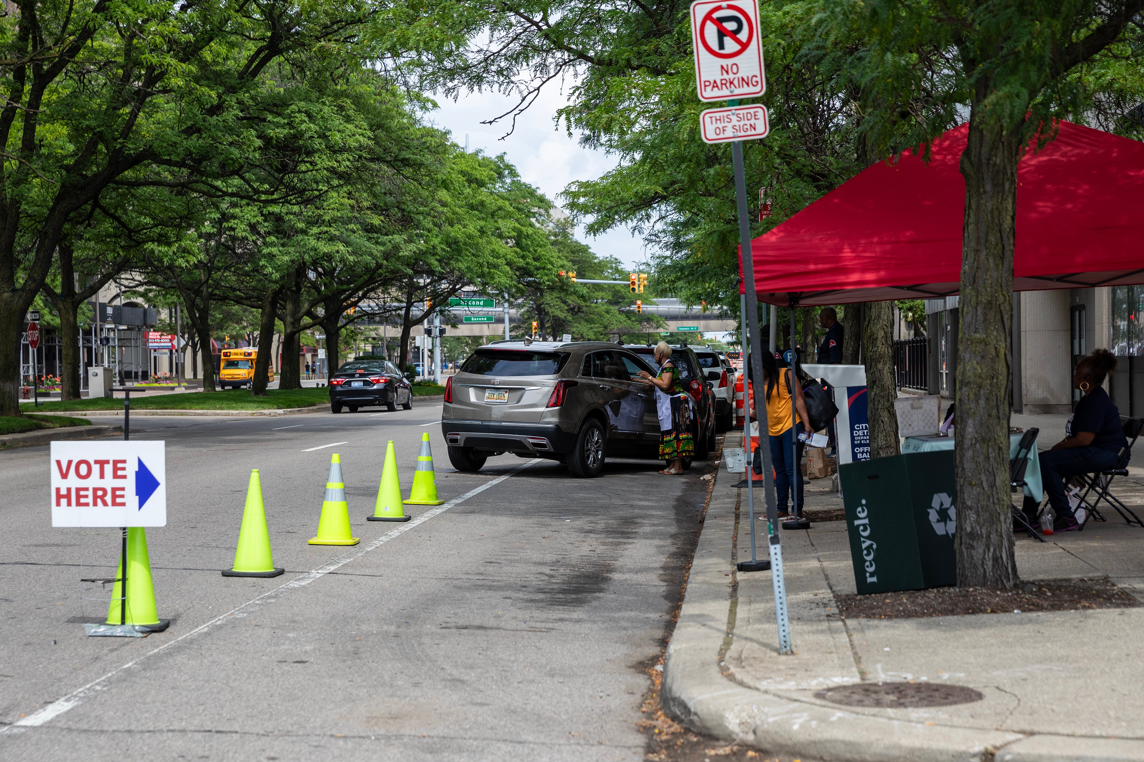 Yellow safety cones lead to a car parked outside of a red tent on the sidewalk near the street.