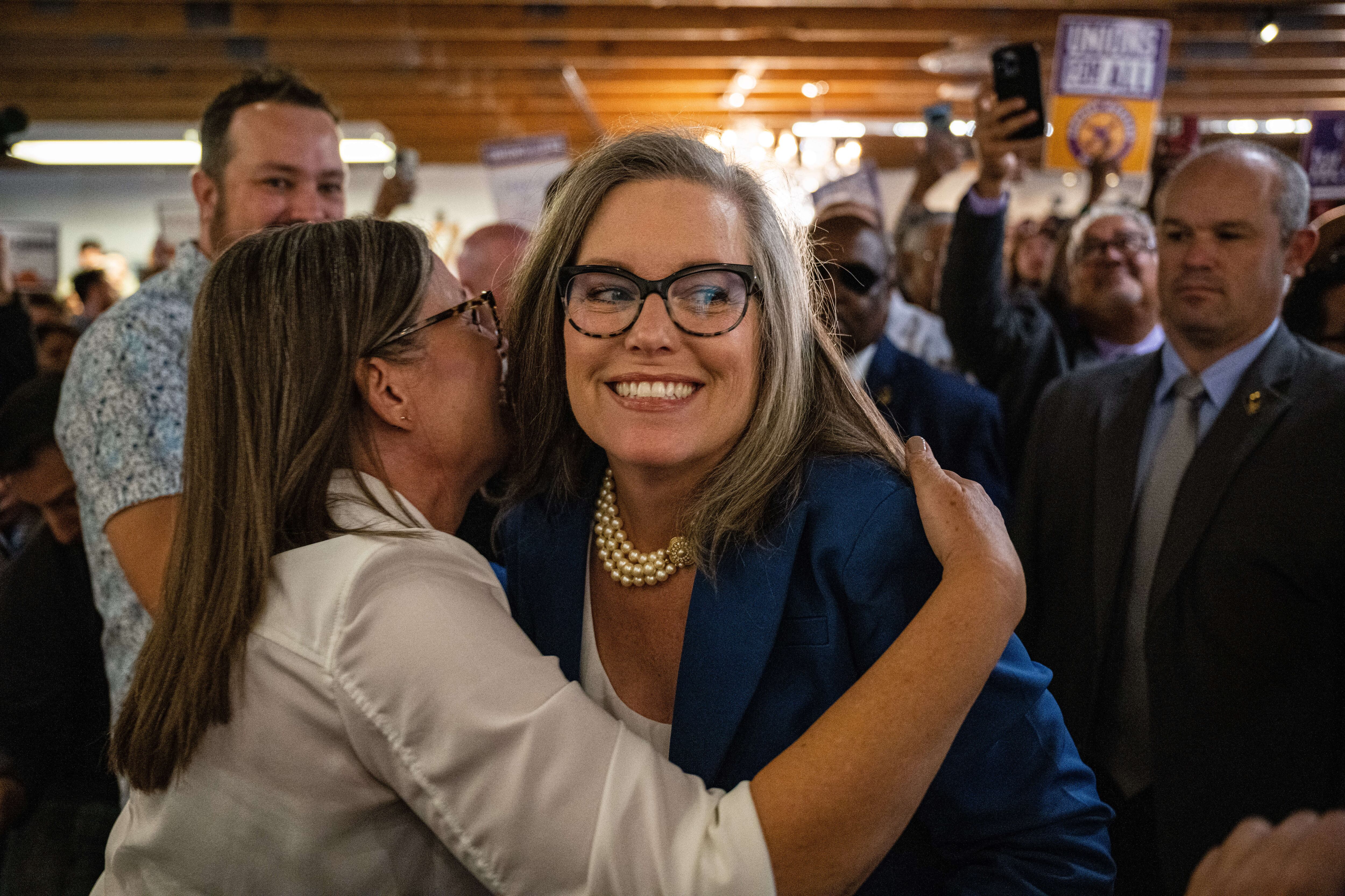 A woman with shoulder-length hair and glasses, wearing a string of pearls, hugs a woman in a white shirt whose back is to the camera.