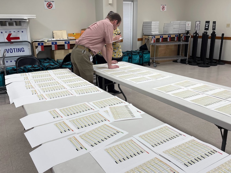 A person stands at the end of a long, white folding table in a room with two folding tables. There are a lot of papers lined up on the tables.