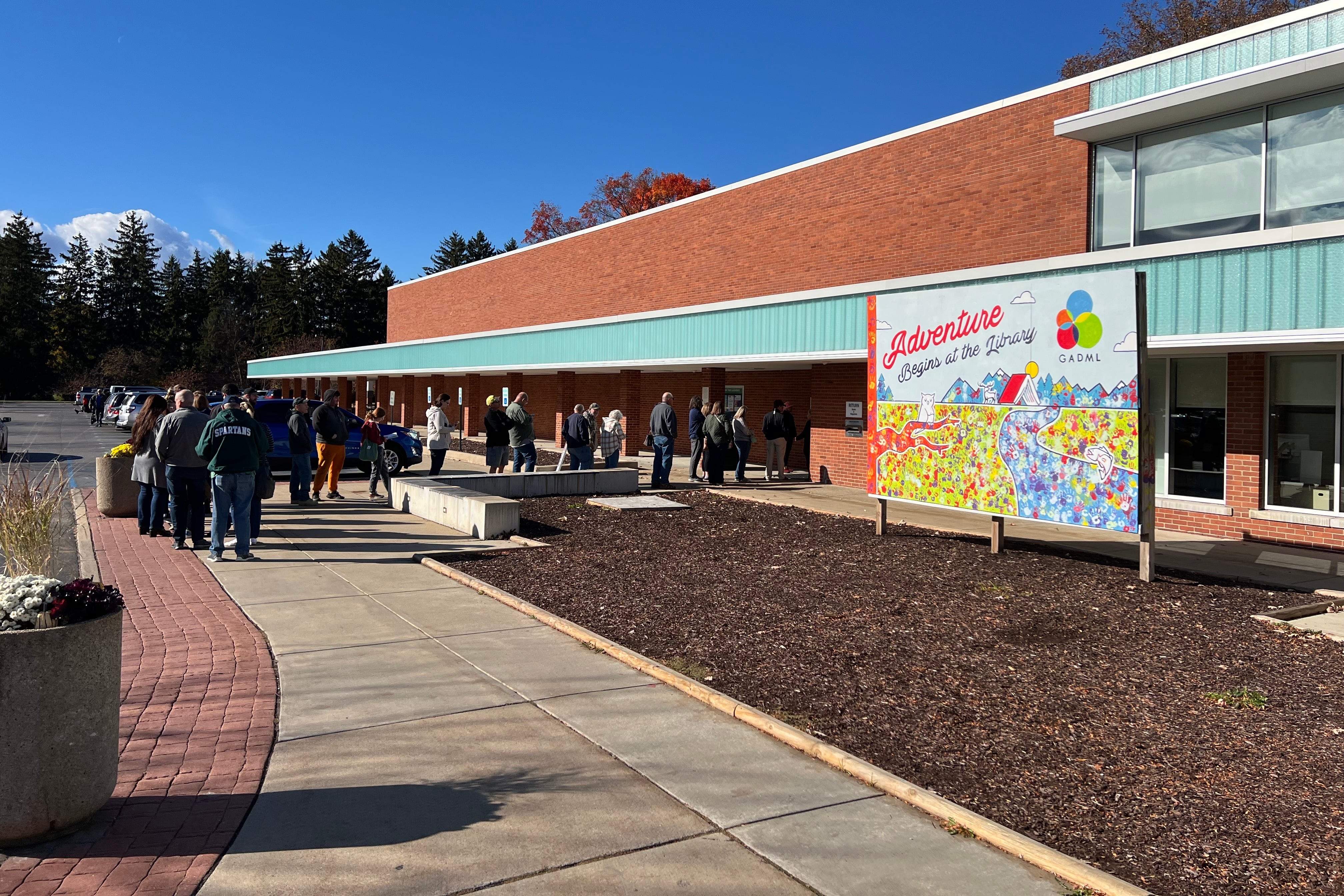 A photograph of a line of people standing outside of a large building on a sunny day.