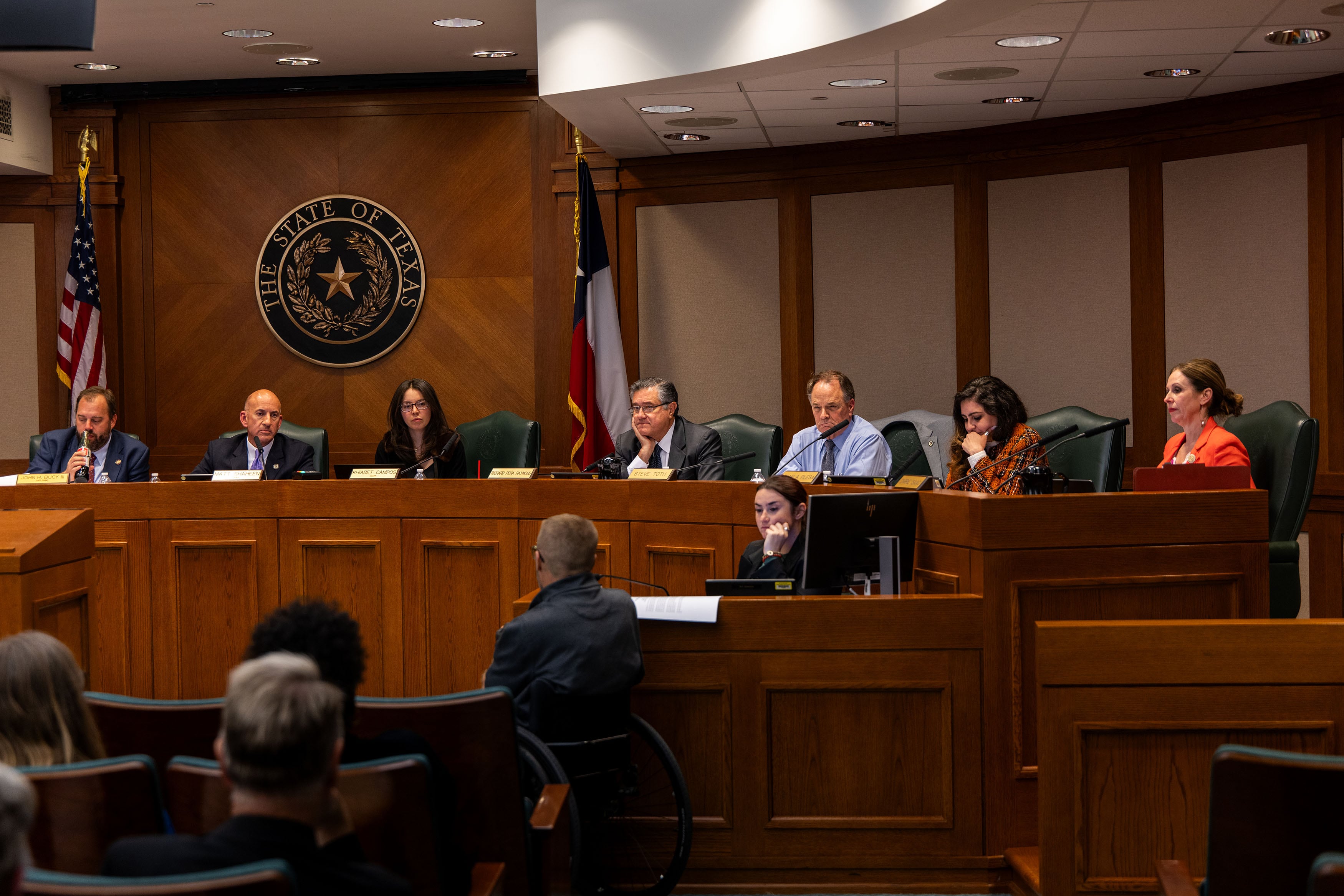 A row of people in business clothes sit in a row in a Capitol building room.