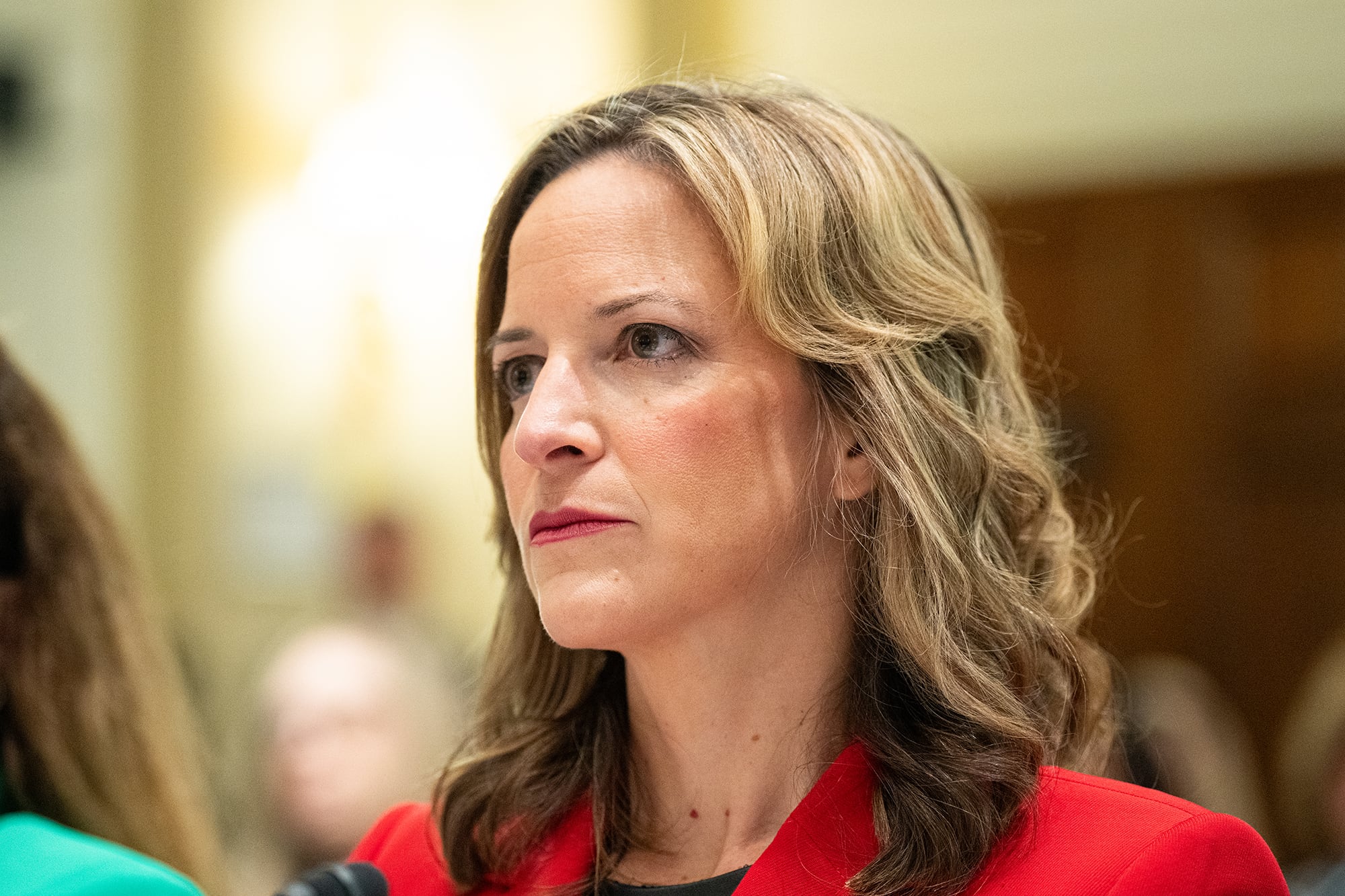A photograph of a white woman with blonde hair and wearing a red suit jacket looks intently in a conference room.