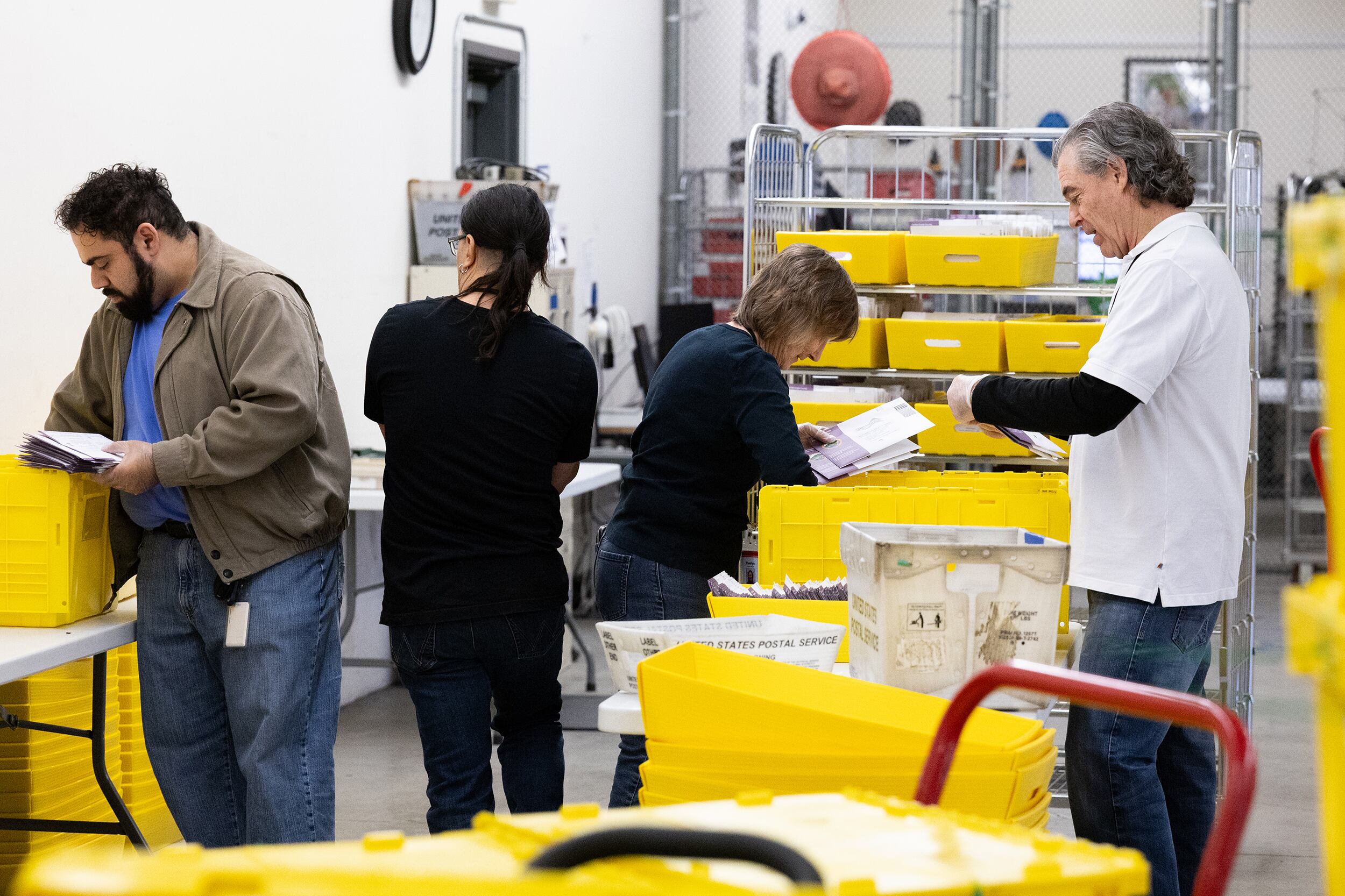 Four people process mail-in ballots in a large room with yellow bins in stacks everywhere.