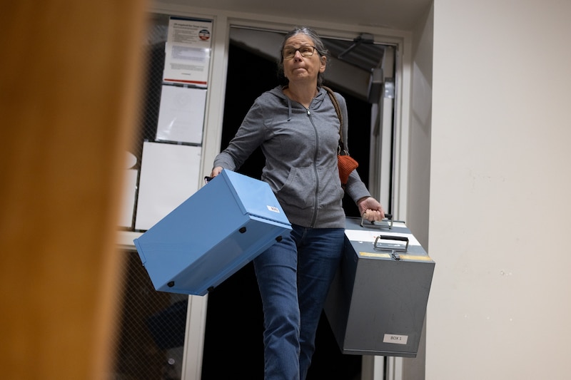 A person wearing a grey sweater and glasses holds two giant boxes with a door way in the background.