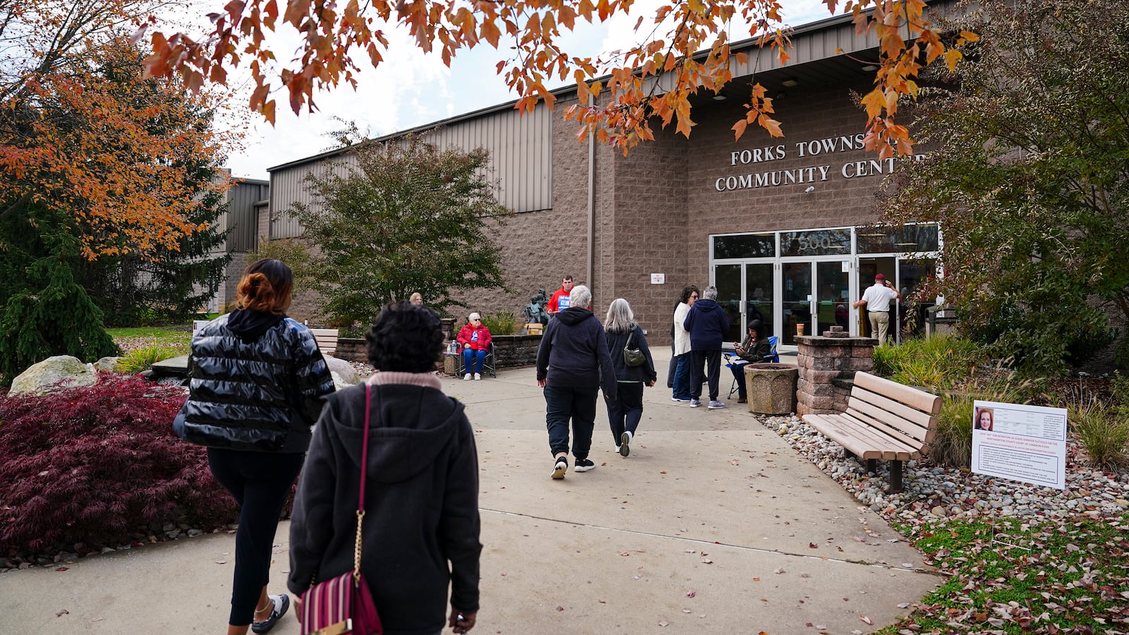 A small crowd of people walk toward the front entrance of a brick community center with fall foliage near the top of the photo.