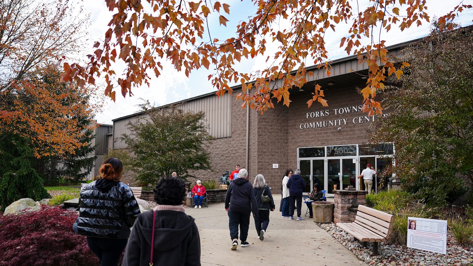 A small crowd of people walk toward the front entrance of a brick community center with fall foliage near the top of the photo.