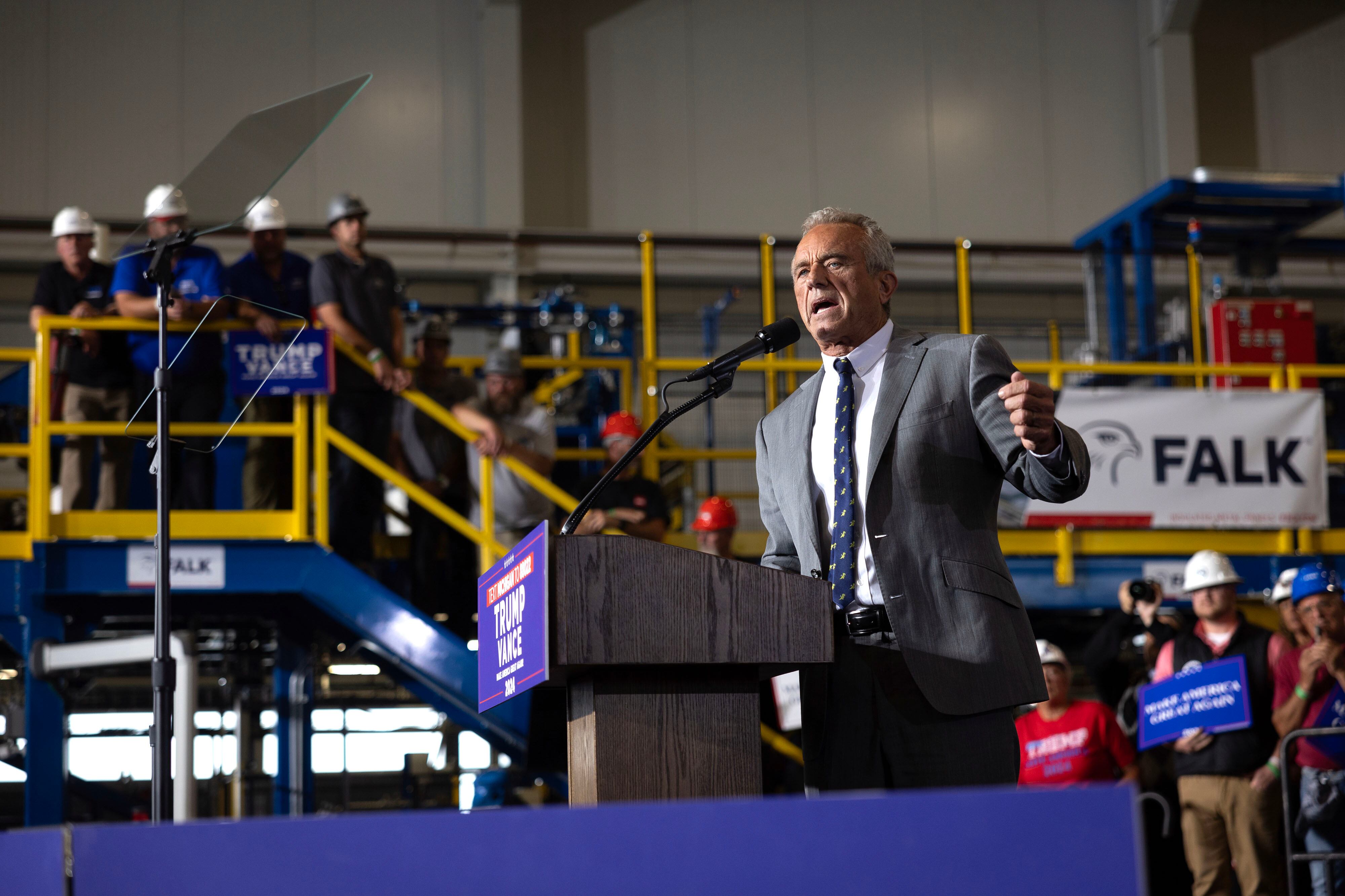 A man in a grey suit stands behind a wooden podium with a microphone with people standing on a walkway in the background.