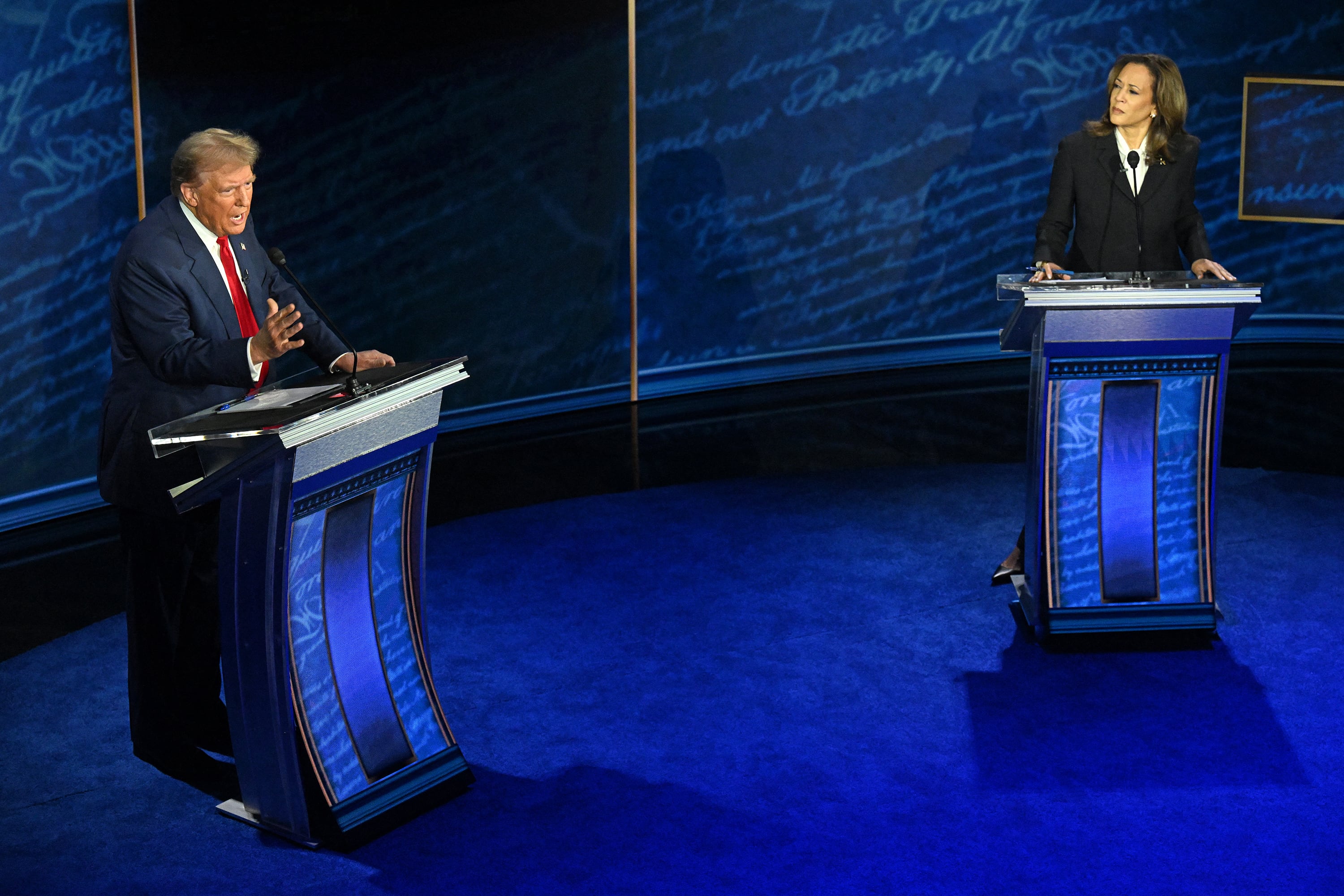 Two adults in suits stand behind blue podiums in a blue room.