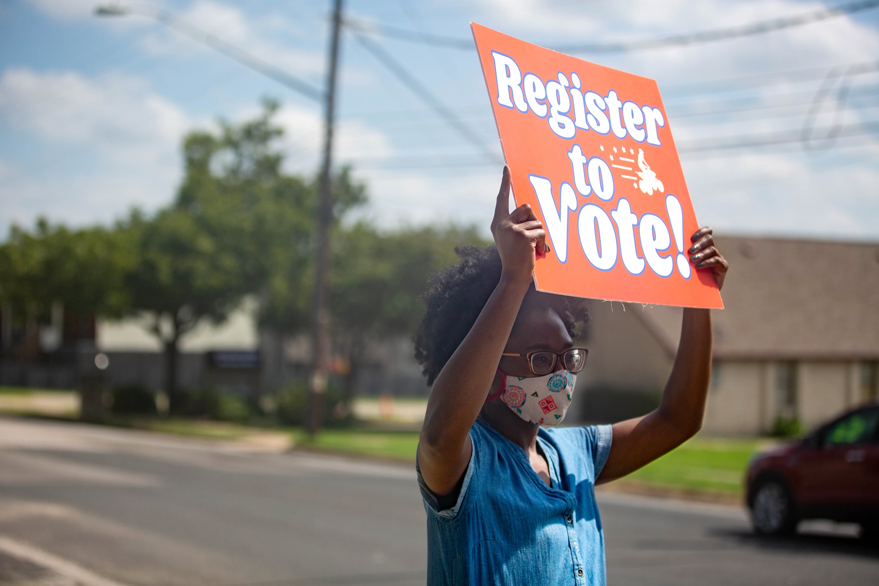 A woman standing on the side of a road holds a sign that says ‘register to vote’
