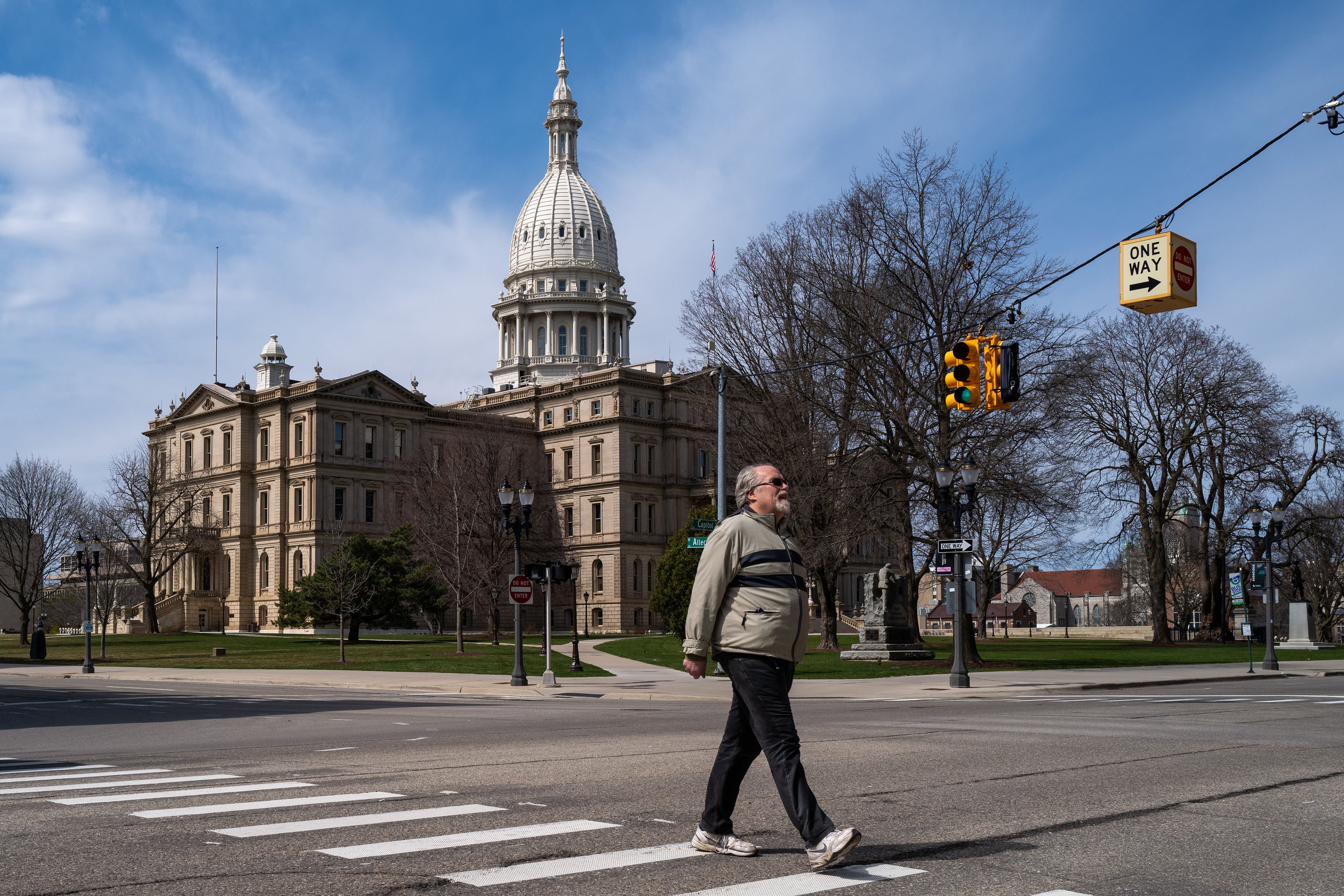 A man in a sweater walks in front of a large stone state capitol building on a sunny day.