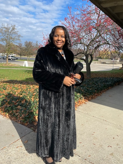 A woman with black hear and a long black dress smiles while posing for a portrait standing in the sun outside with a blue sky and a tree in the background.