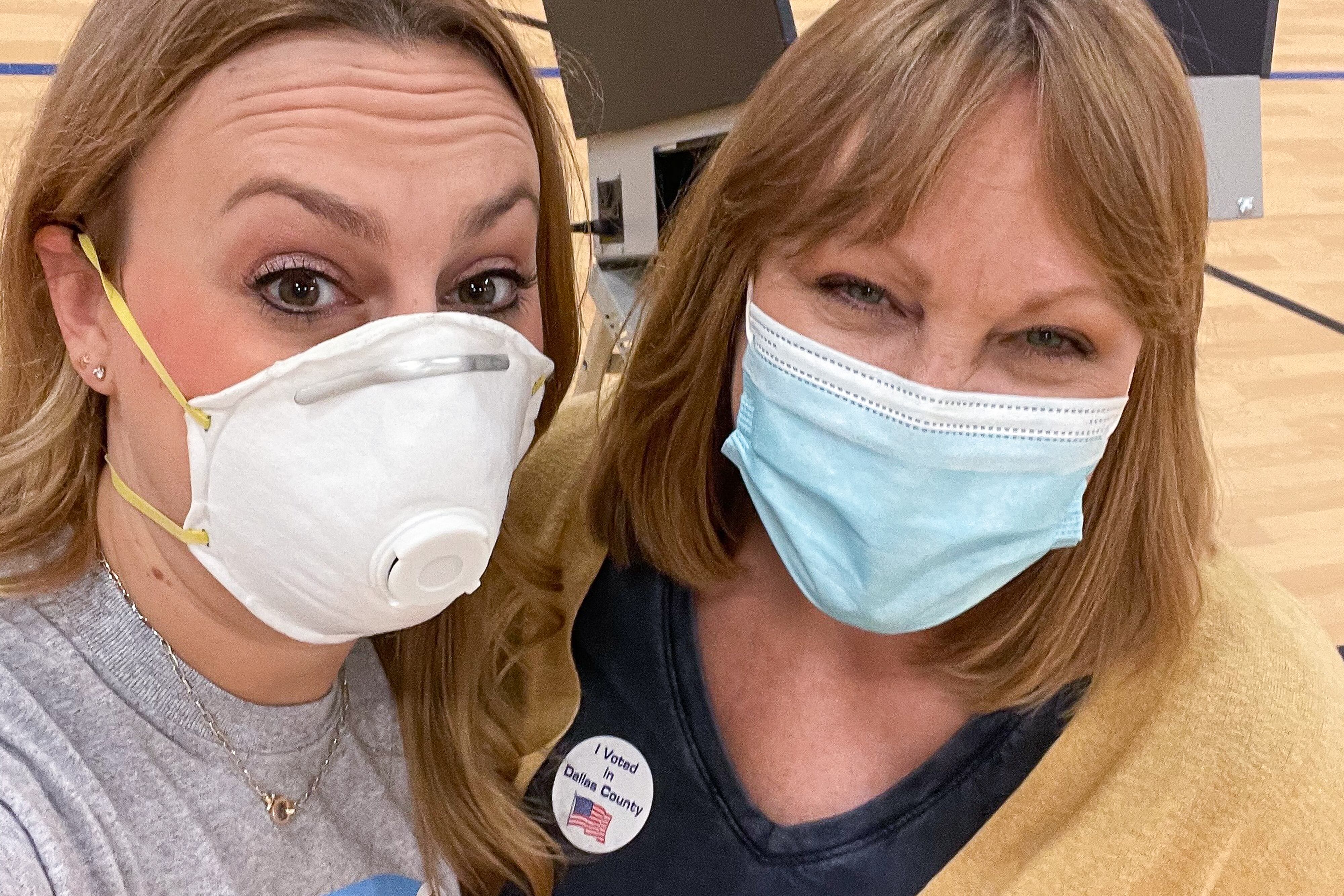 Two women wearing face masks pose inside a gymnasium with voting booths behind them.