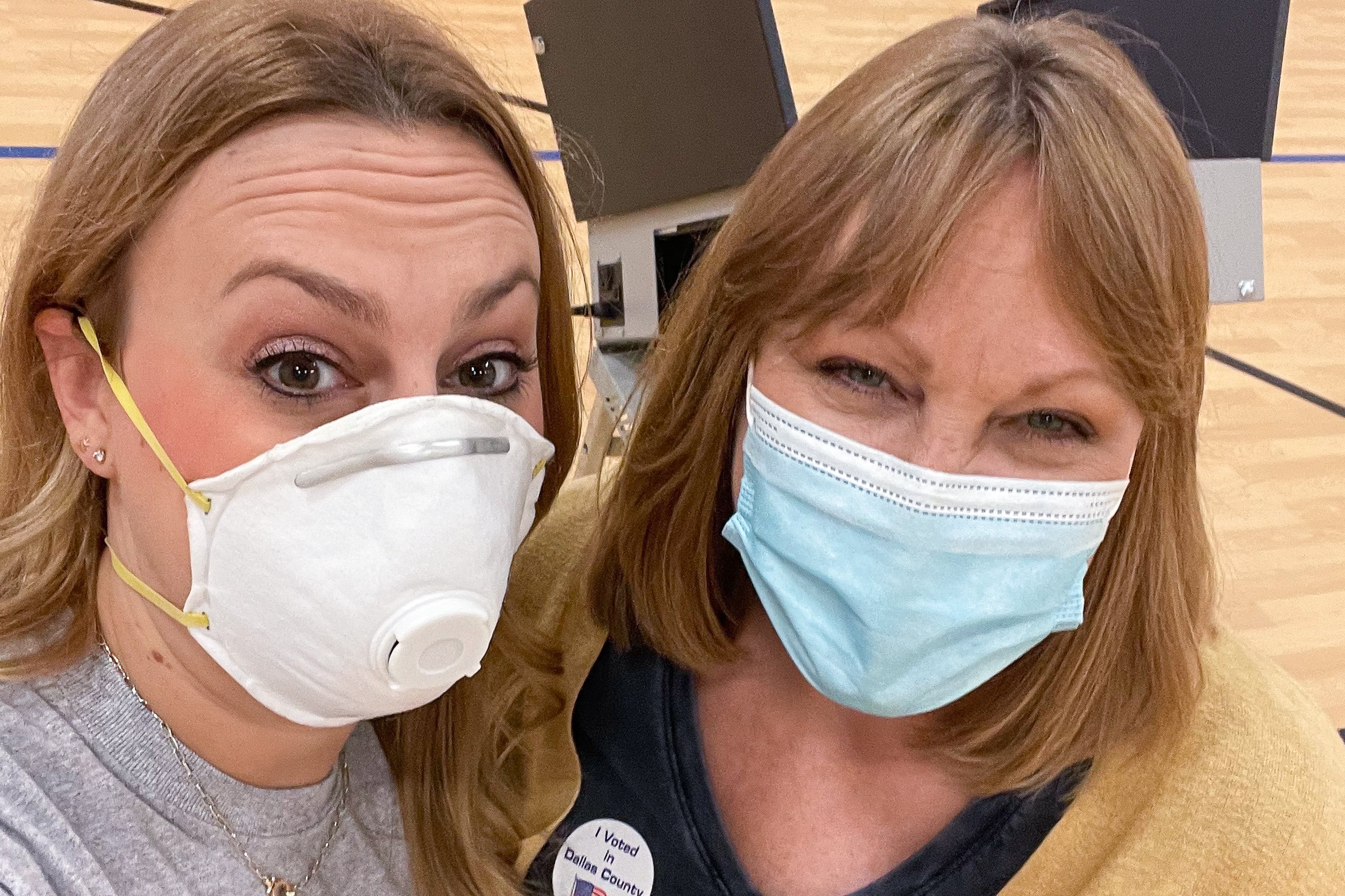Two women wearing face masks pose inside a gymnasium with voting booths behind them.