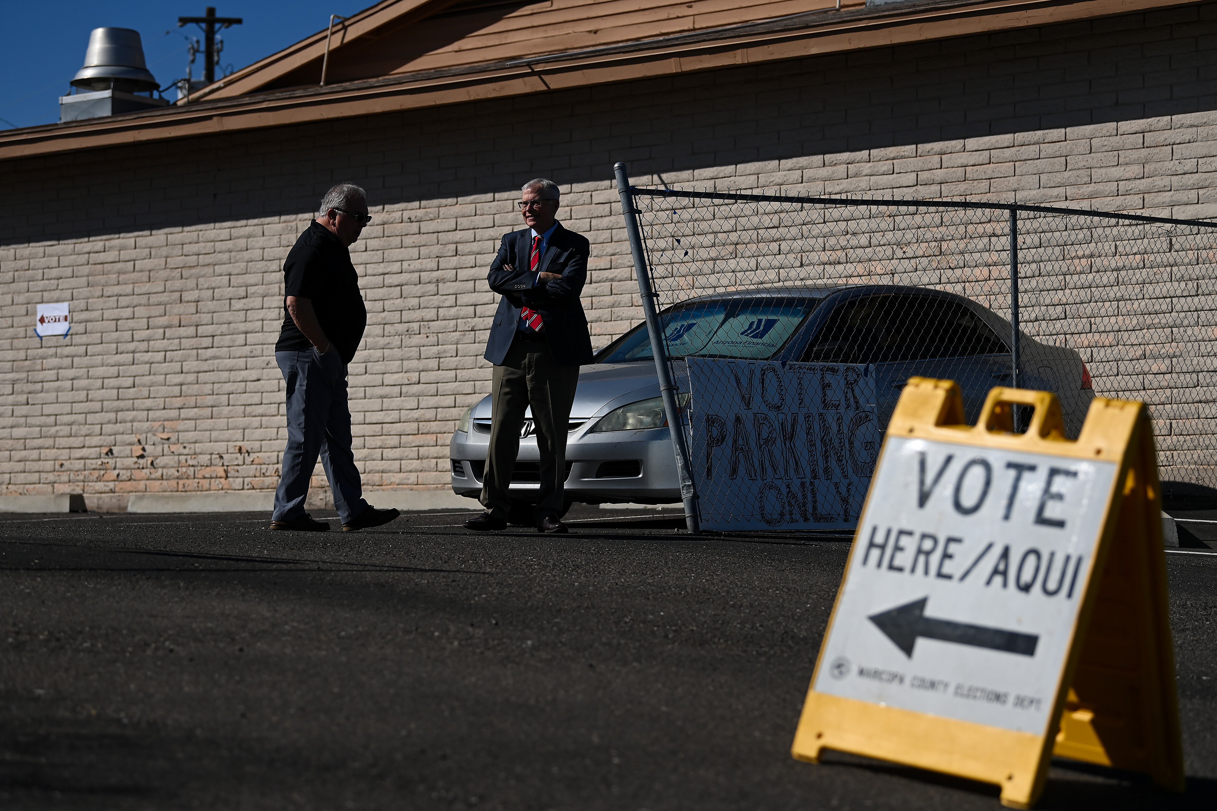 Two men stand outside a brick building behind a prominent yellow sign reading "Vote Here / Aqui"