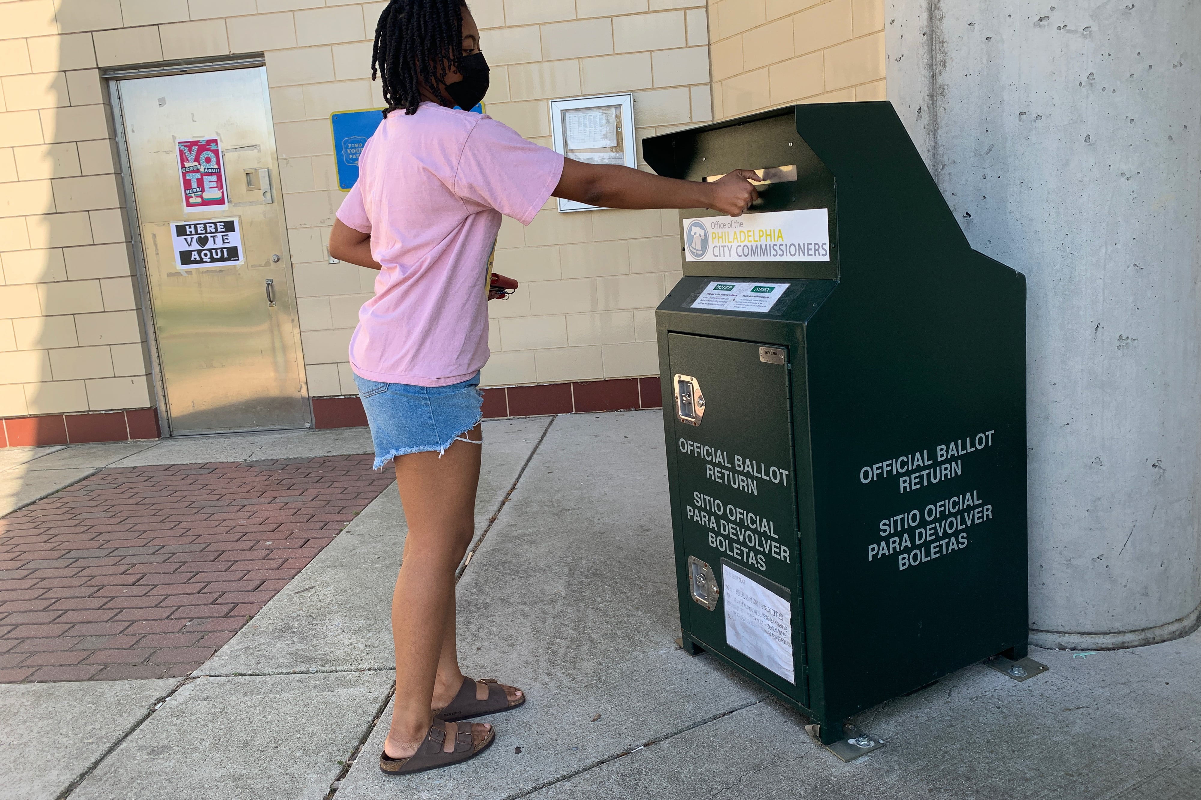 A woman in a pink shirt and shorts puts a ballot through the slot of a ballot dropbox.
