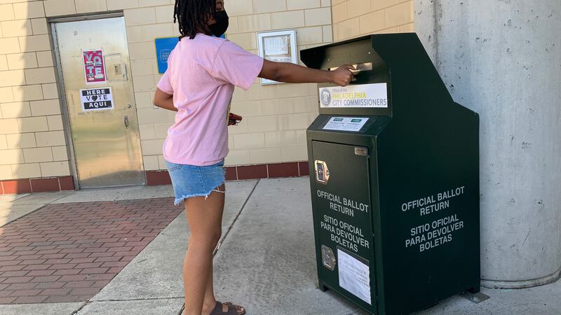 A woman in a pink shirt and shorts puts a ballot through the slot of a ballot dropbox.