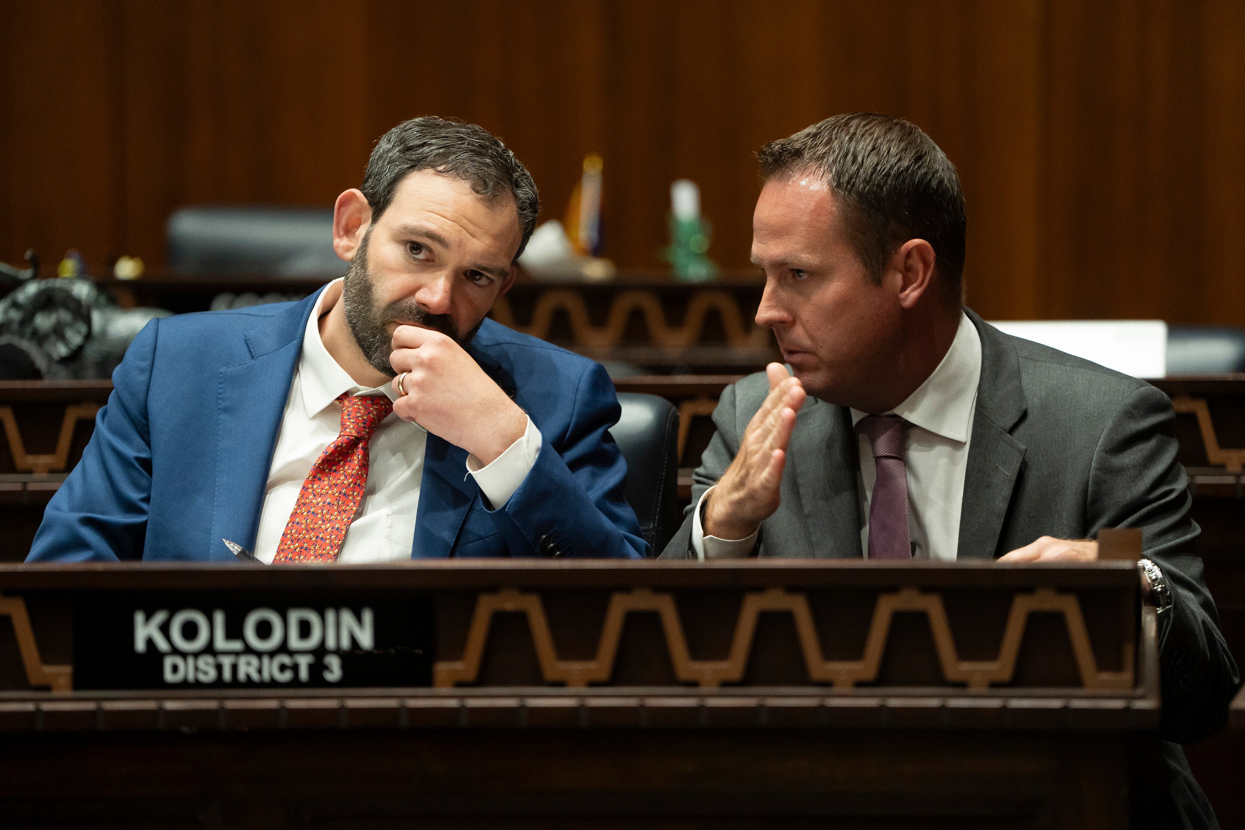 Two men in suits speak to each other while sitting at a wooden desk.