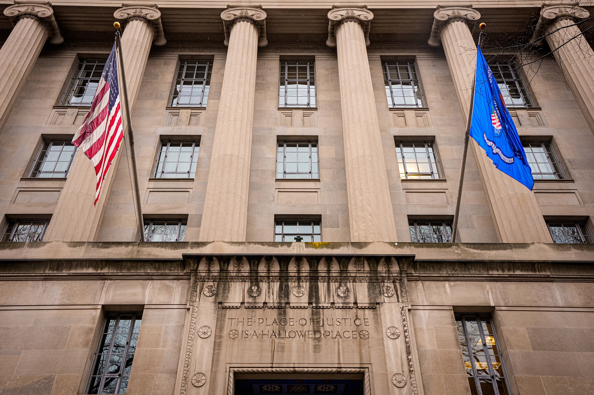 A photograph of an entrance to a large stone building with two flags on each side of the doors.