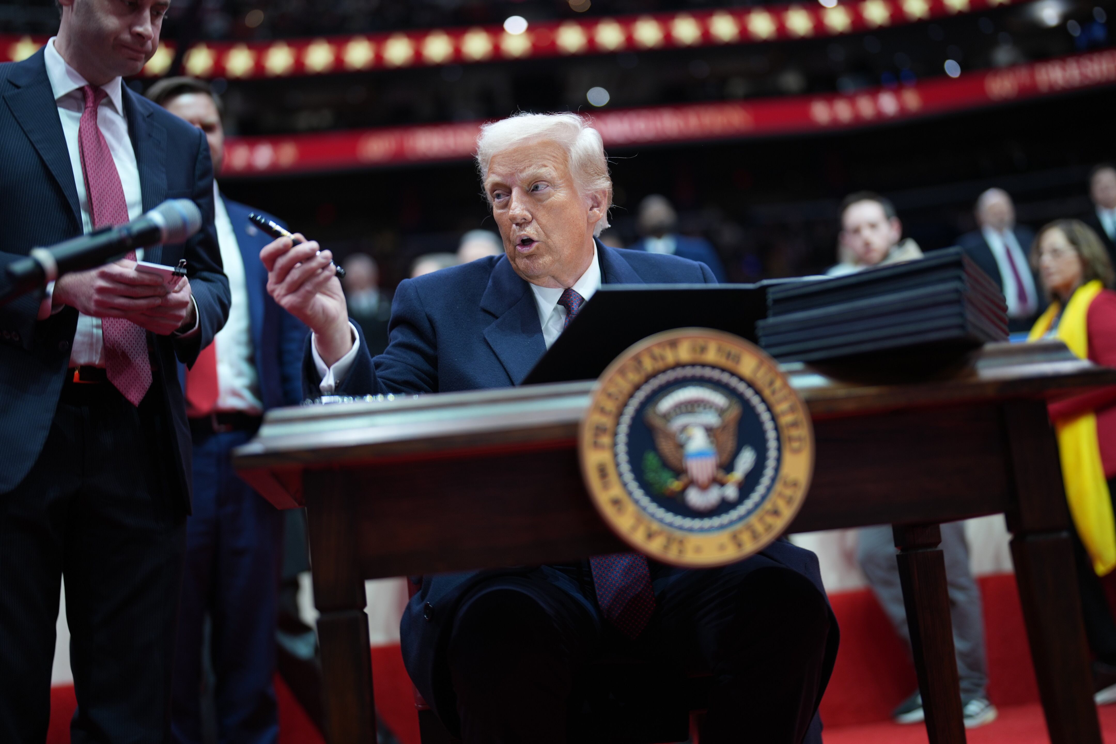 A man with white hair and wearing a suit holds a pen and sits at a desk in a stadium.