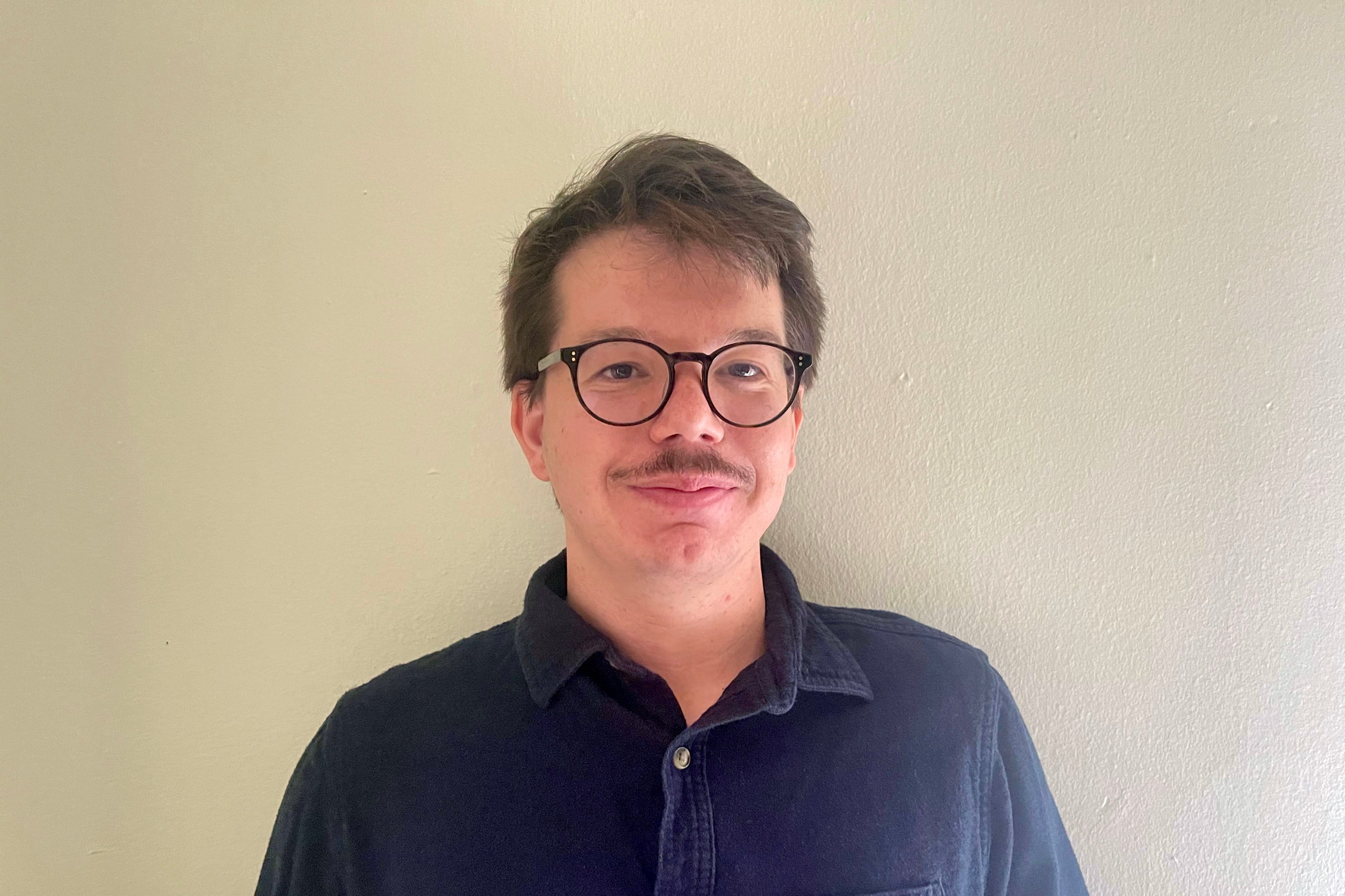 A man with short dark hair, wearing black glasses and a blue dress shirt poses for a portrait in front of a tan wall.