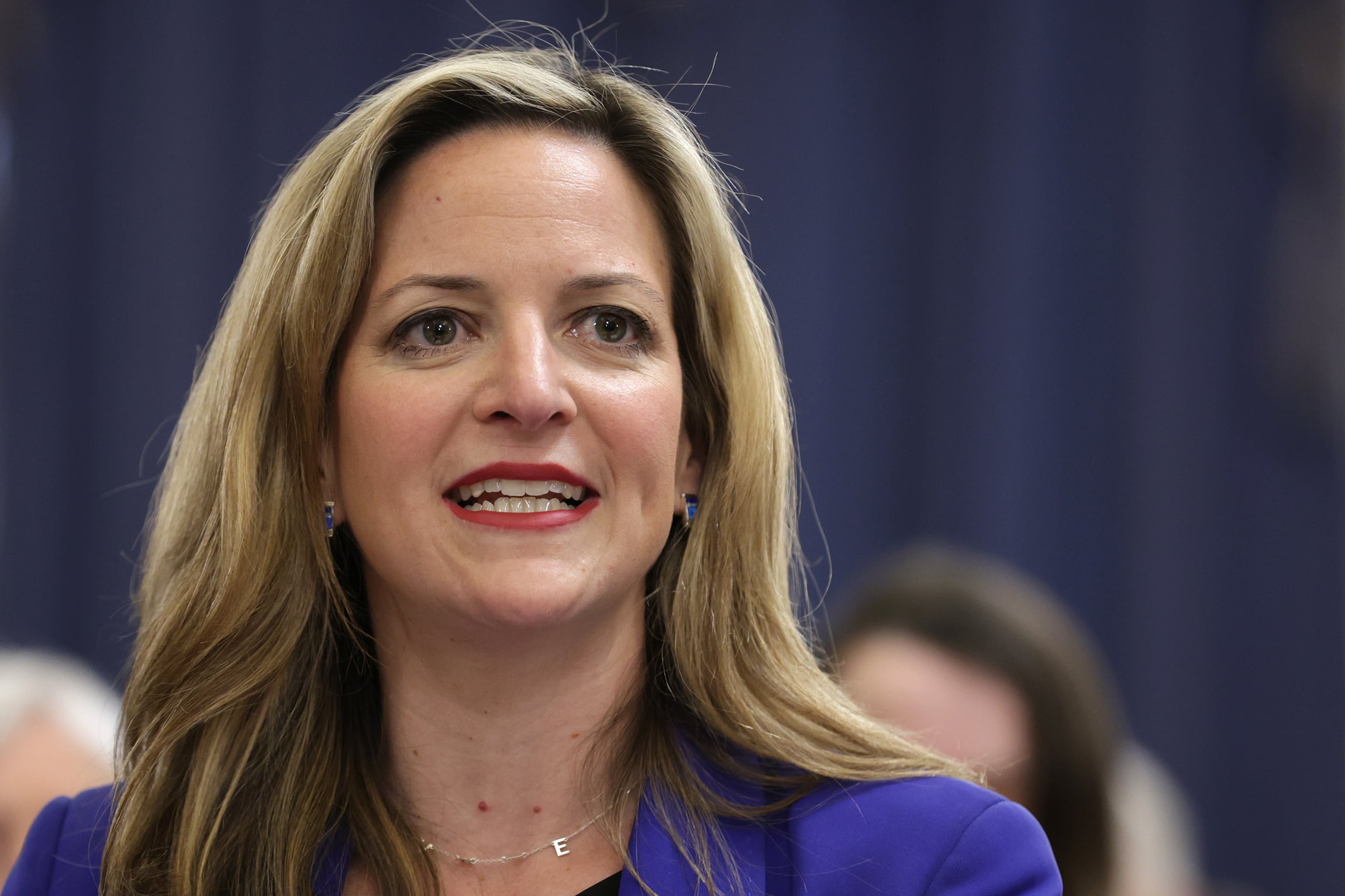 A photograph of a white woman with long blonde hair talking in a room with other people in the background.