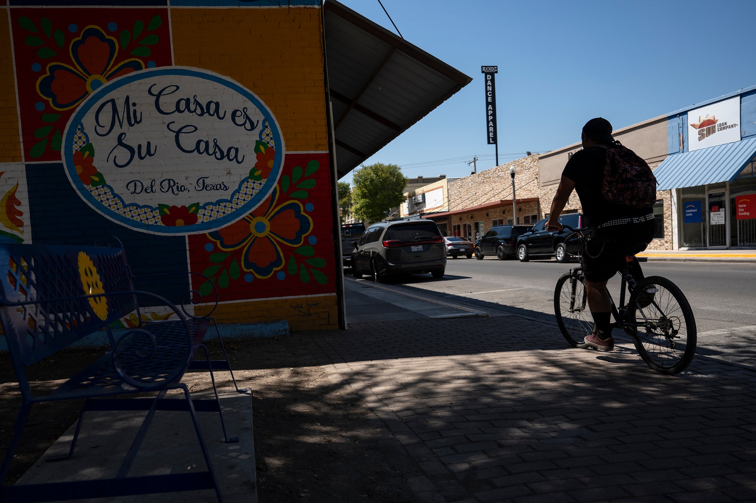 A photograph of a person riding a bike outside on a small downtown street next to a building that has a mural that reads "Mi casa es su casa."