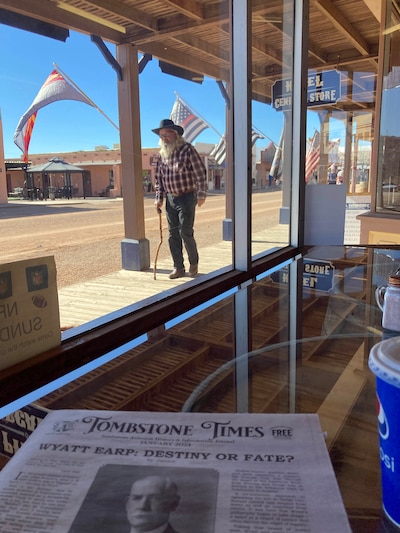 A man with a long white beard and wearing a hat and using a walking cane walks down a wooden sidewalk with flags in the background and a newspaper on a table in the foreground.