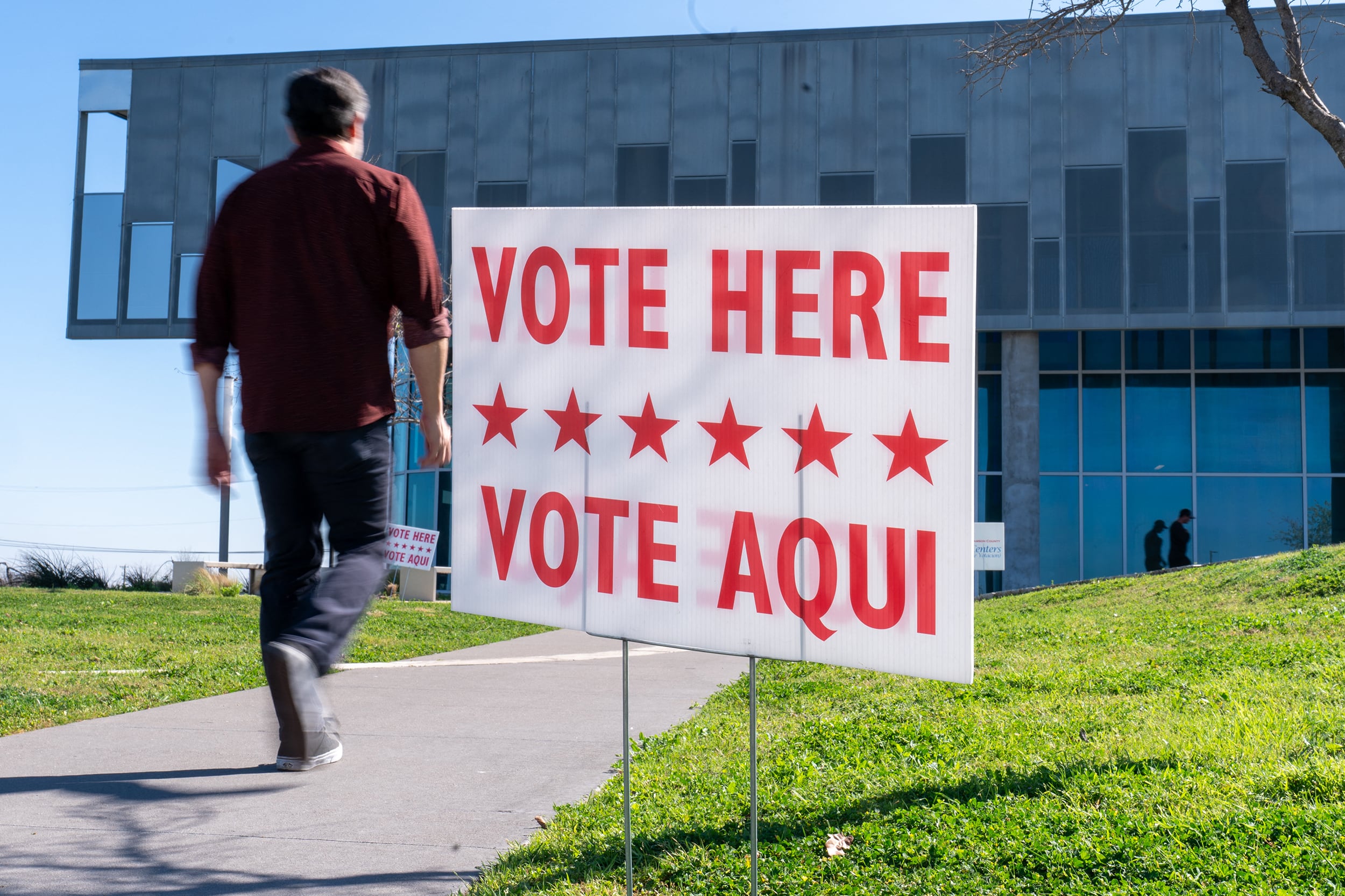 A person walks by a sign that reads "Vote Here Vote Aqui" outside of a large glass building on a sunny day.