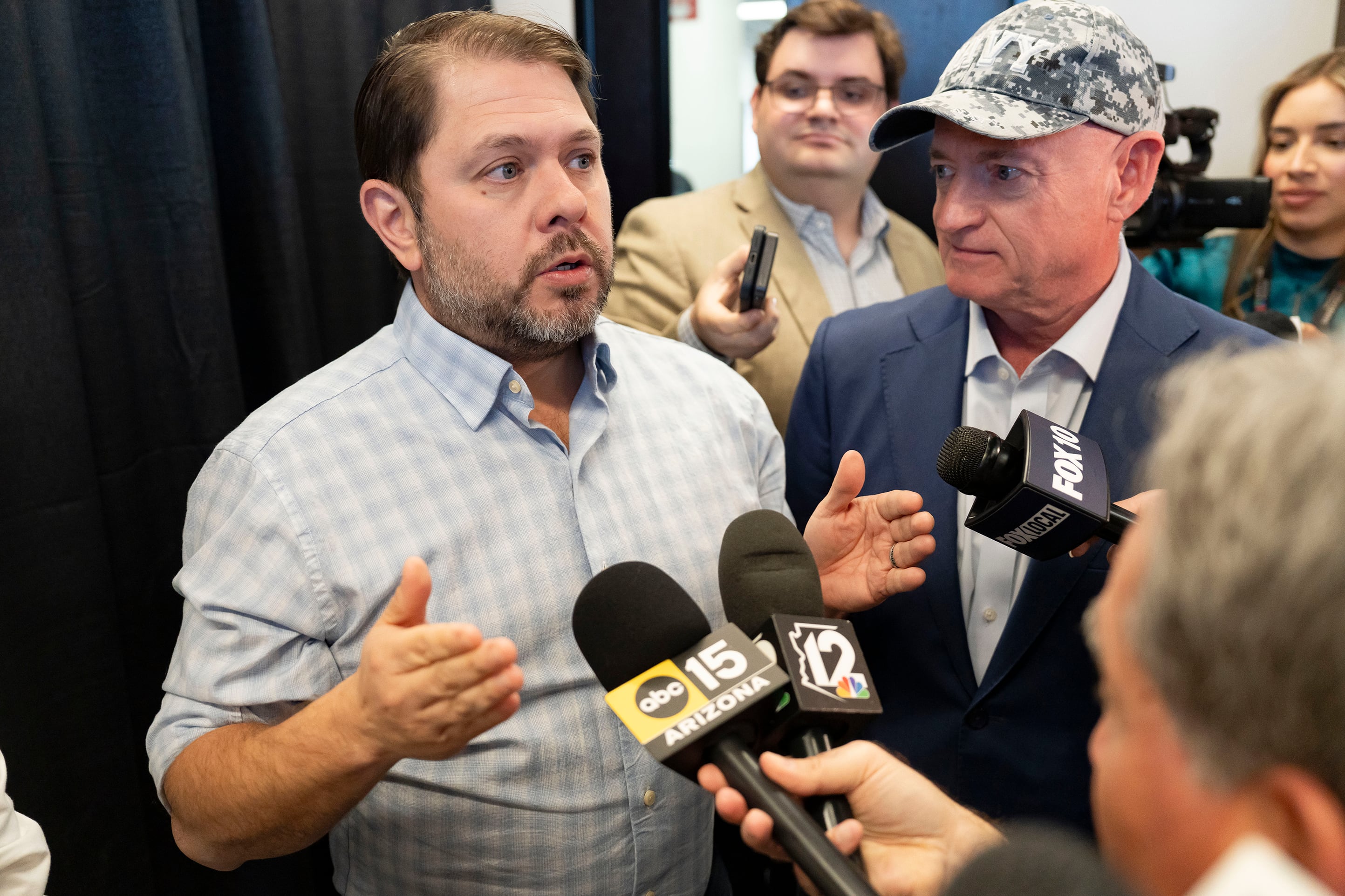 A photograph of two men standing in a crowd of reporters speaking into microphones.