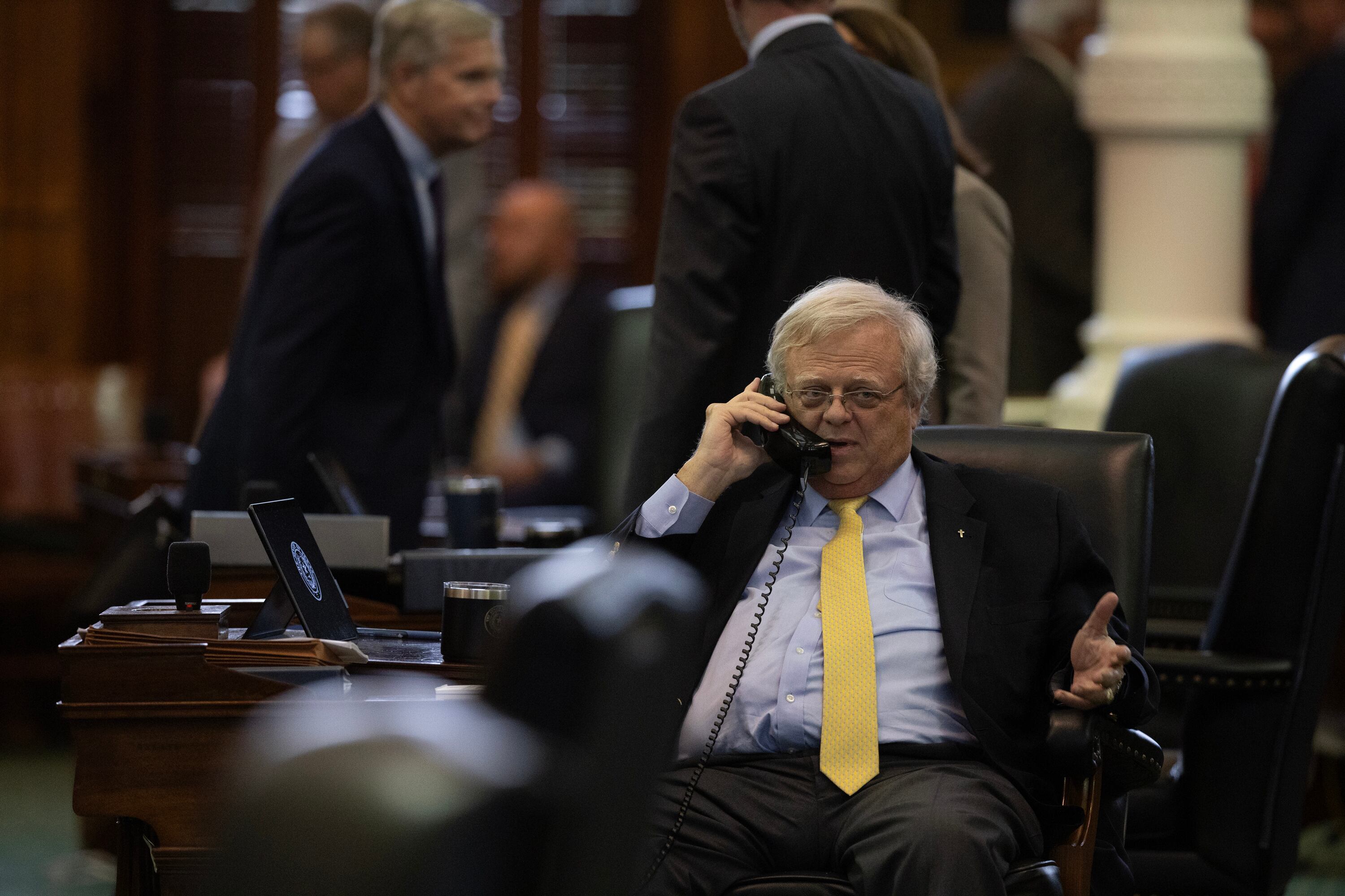 A man in a suit jacket, light blue shirt, and yellow tie talks on the phone.