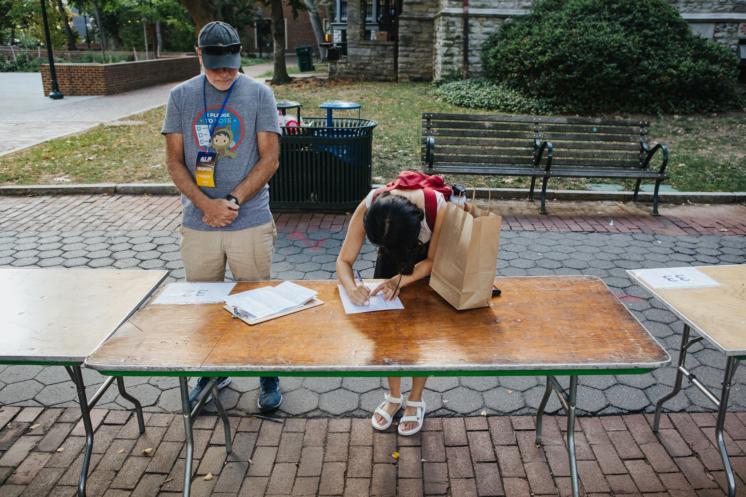 Two people stand next to a wooden school table that's outside on a brick pathway.
