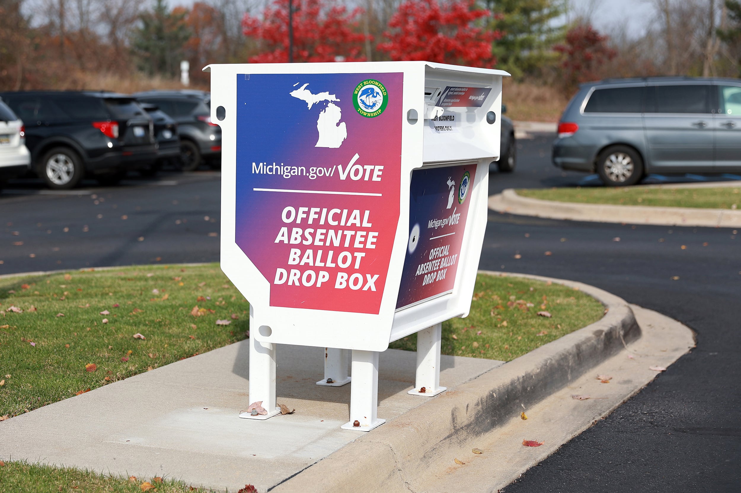 A red, white and blue drop box for ballots sits on the side of a parking lot on a sunny day.