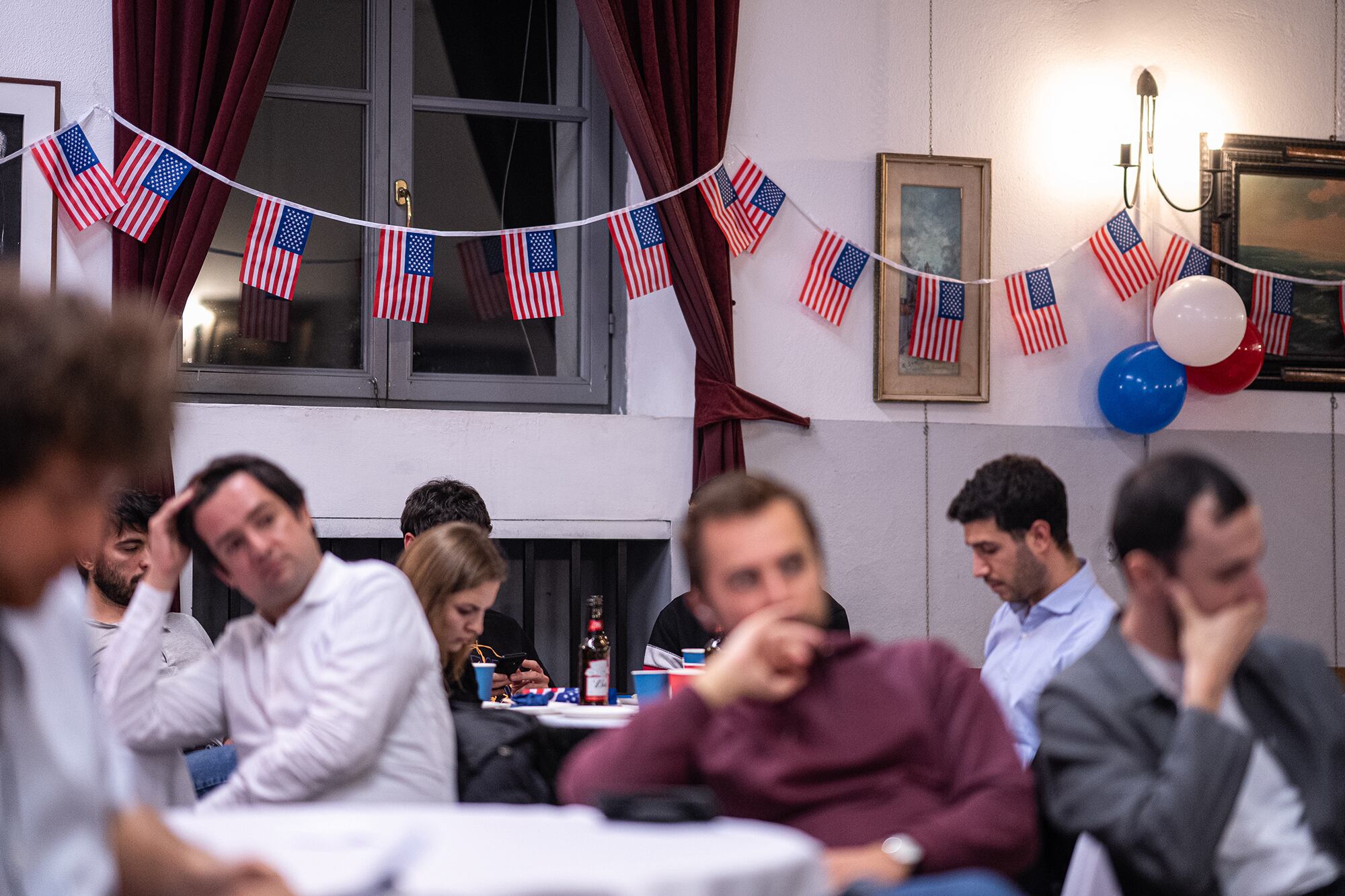 A photograph of a group of men sitting at tables in a room with a string of American flags in the background wall.