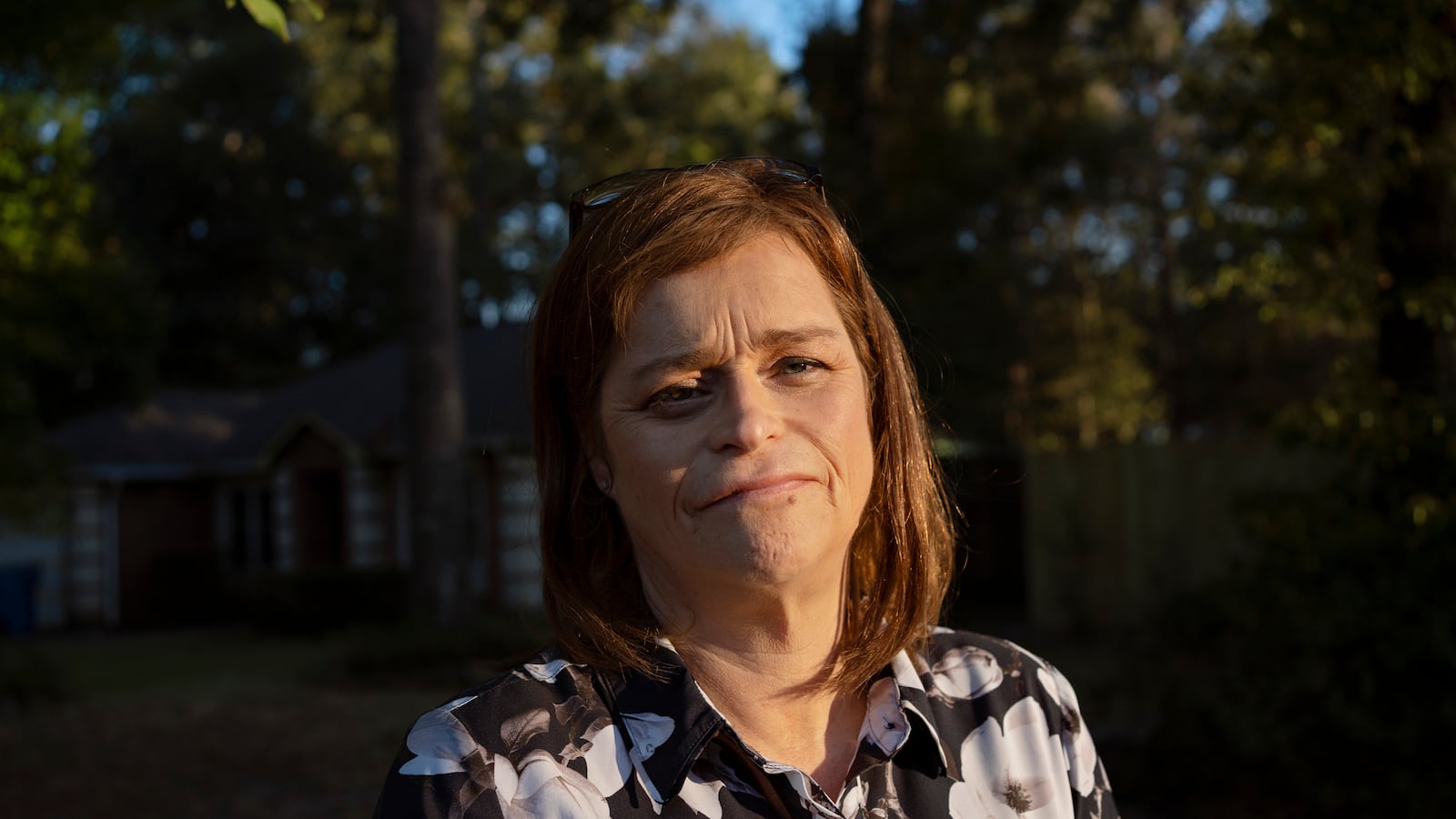 A woman with short brown hair and wearing a floral blouse stands in front of a bunch of trees and poses for a portrait.