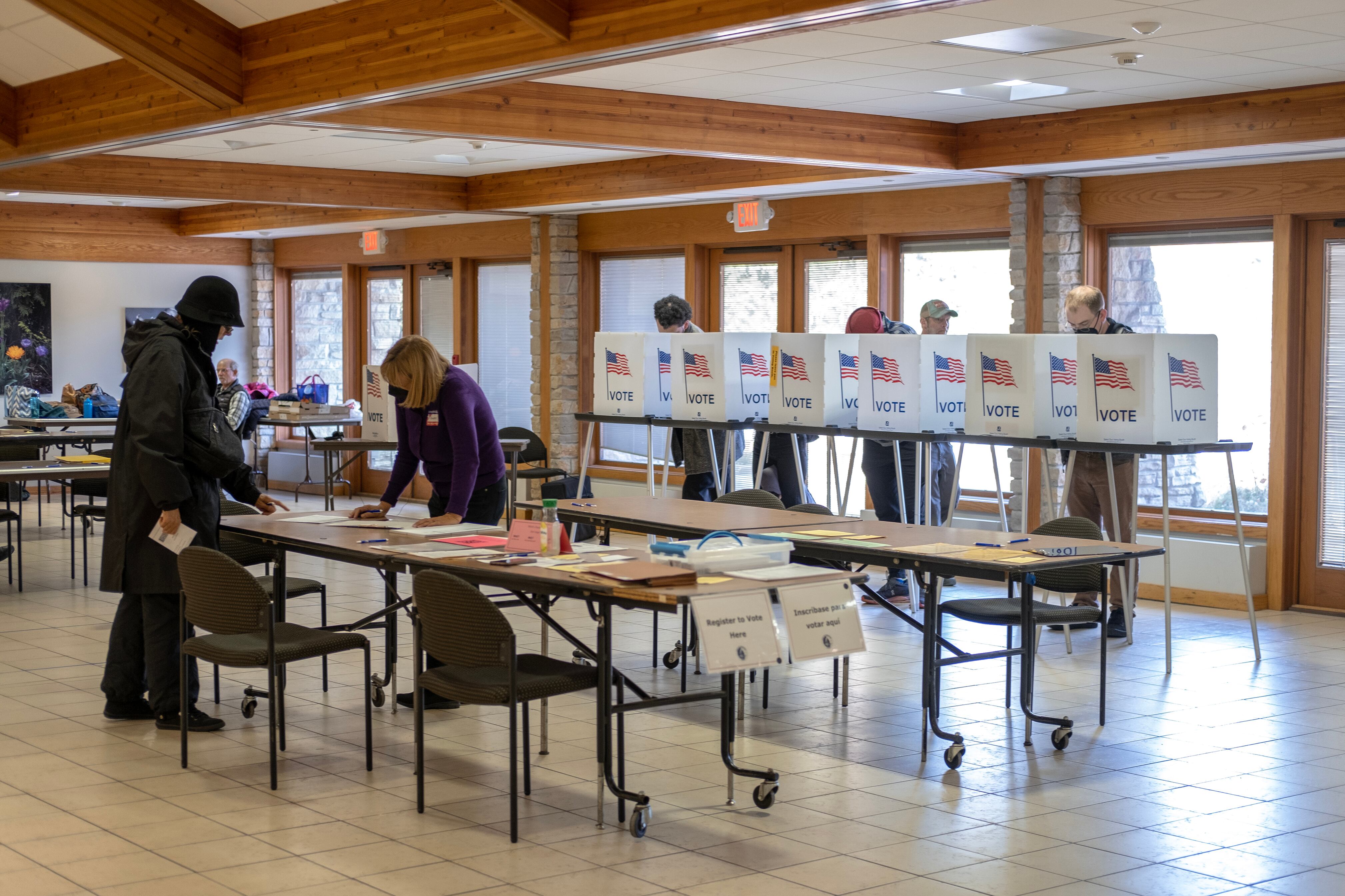 Two people talk over a table while four people vote in the background.