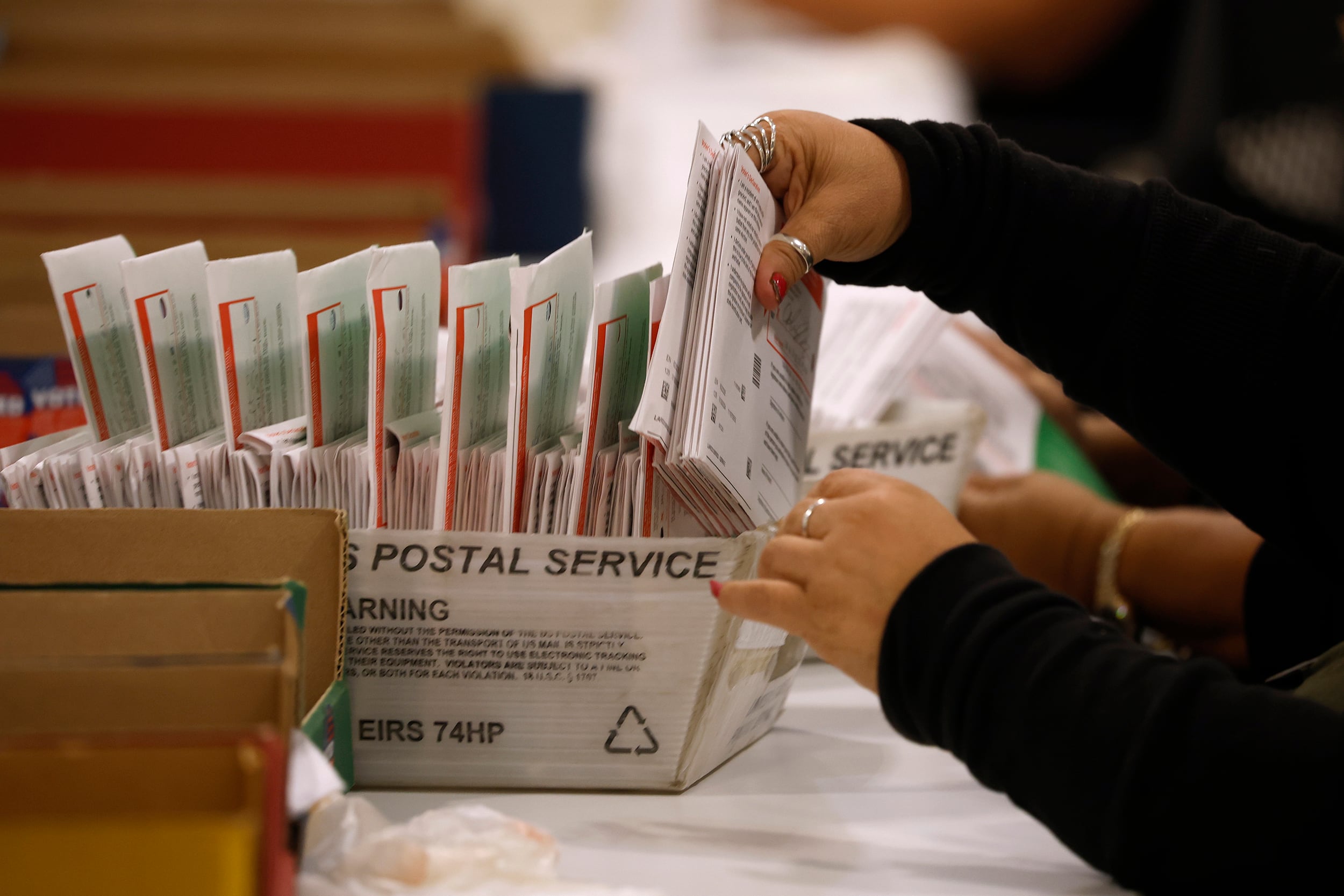 A photograph of a pair of hands working with mail in ballots in a USPS sorting box.