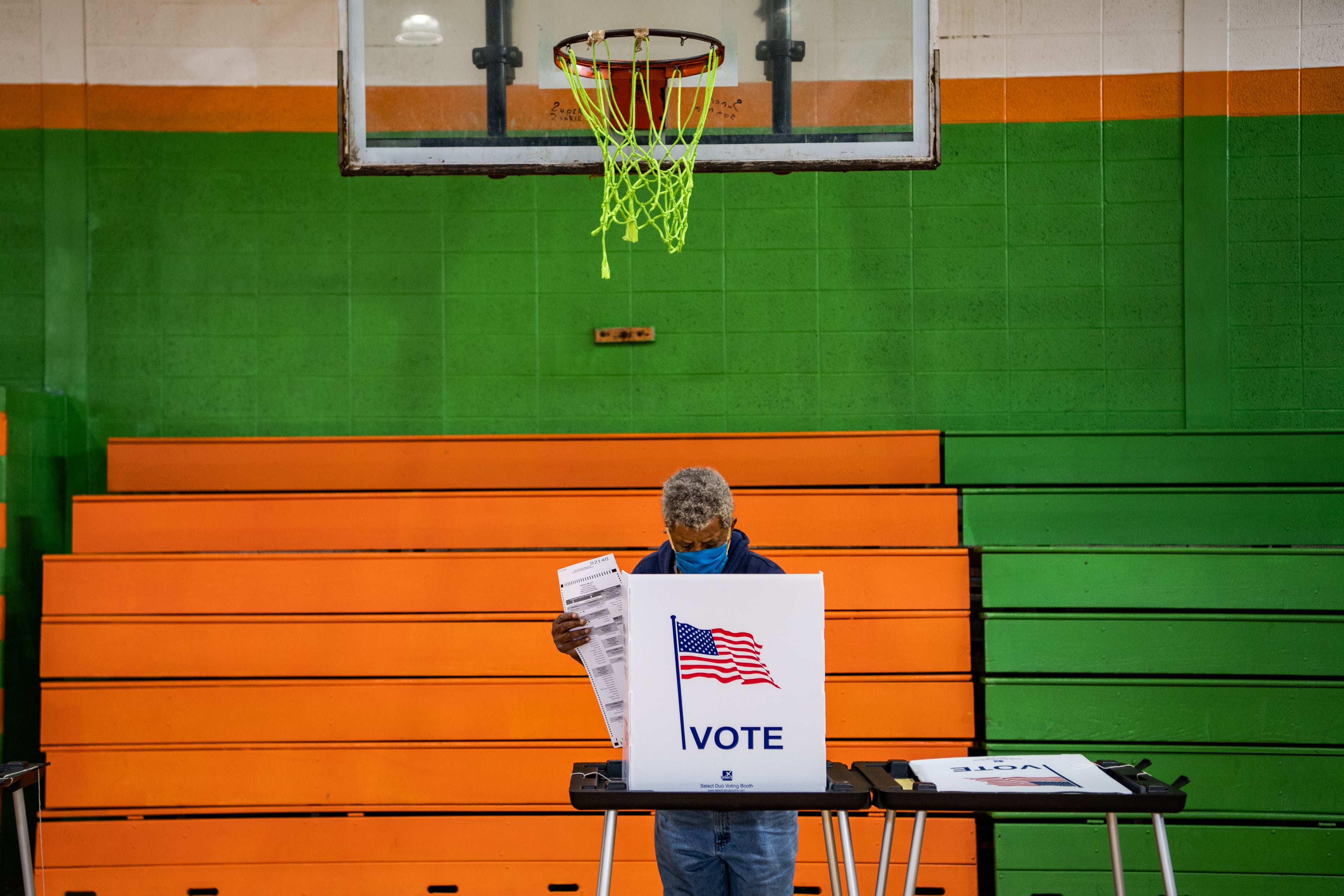 A man, wearing a blue surgical mask, votes at a polling station on a basketball court. The walls and bleachers behind him are bright green and orange.