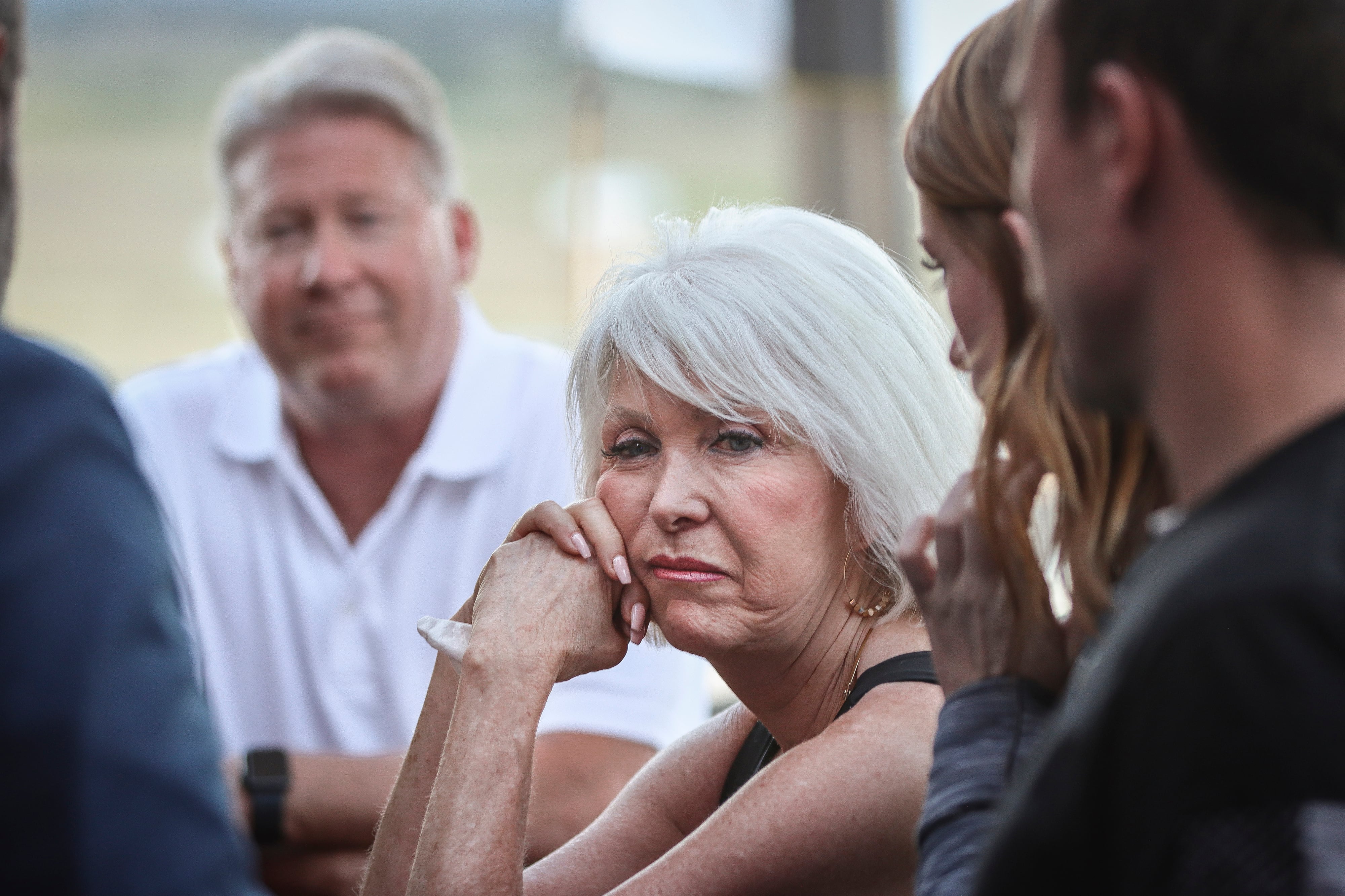 A woman with short white hair holds her face up with her hands while looking off between a group of people. There's a man in a white shirt in the background.