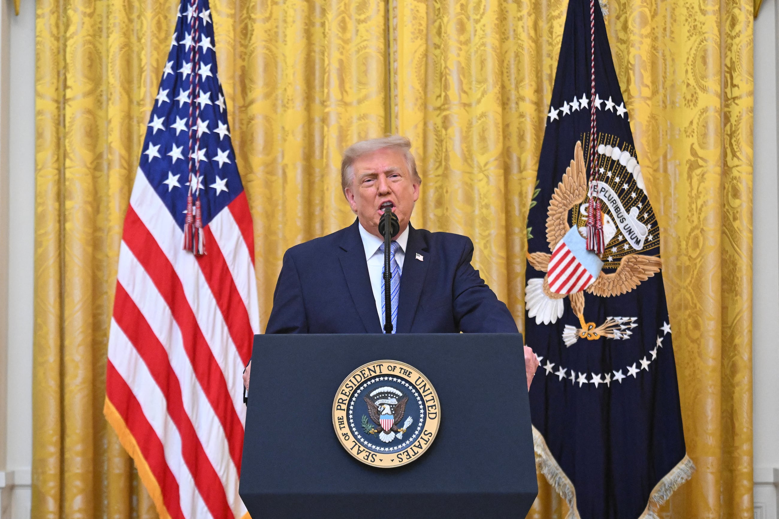 Donald Trump speaks from a podium in front of two flags and a yellow background.