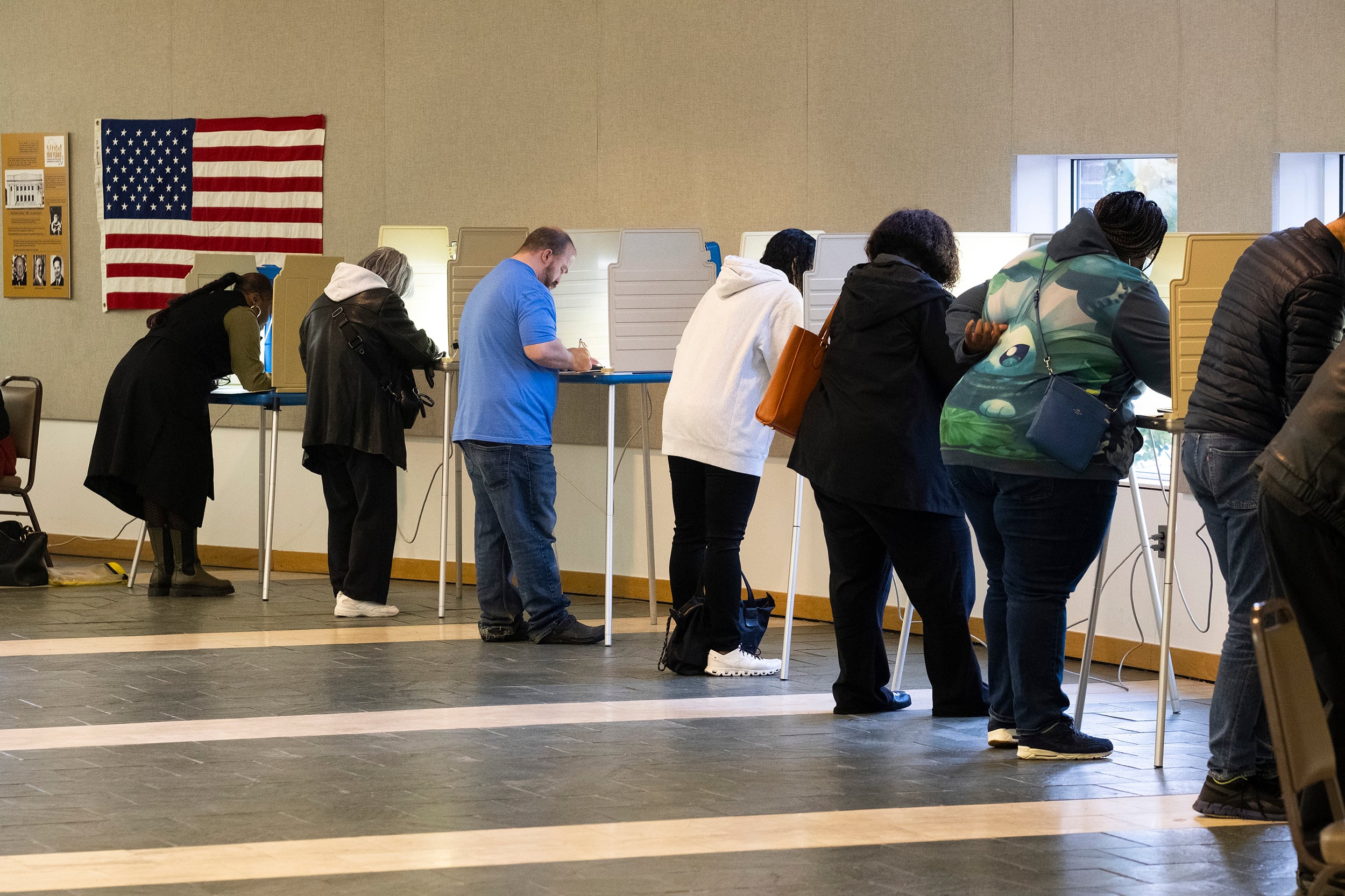A line of people stand near voting boxes where they fill out their ballots in a room with tan walls and an American flag in the background.