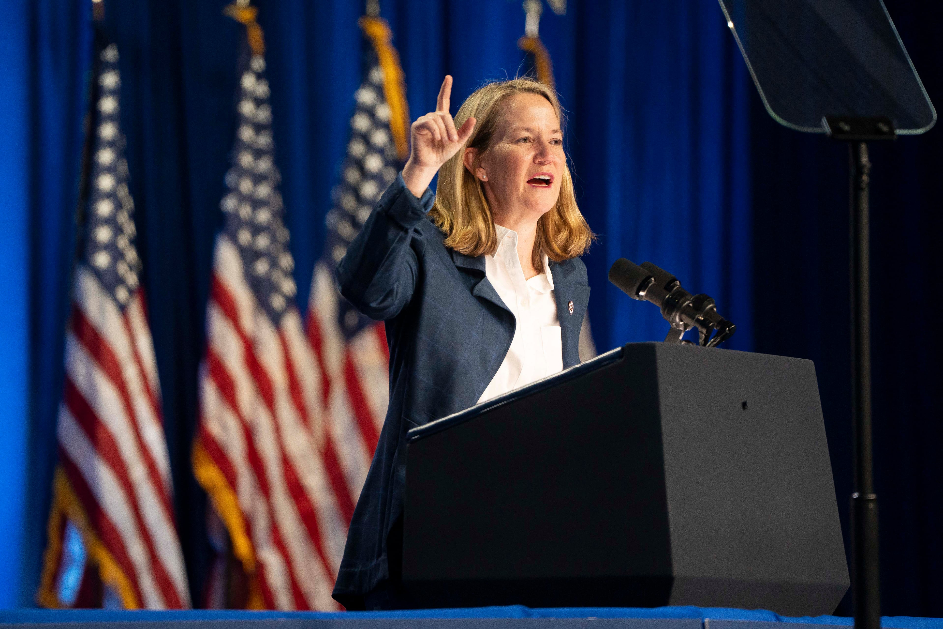 A white woman with blonde hair speaks from behind a podium with her right hand in the air and there are three American flags in the background.