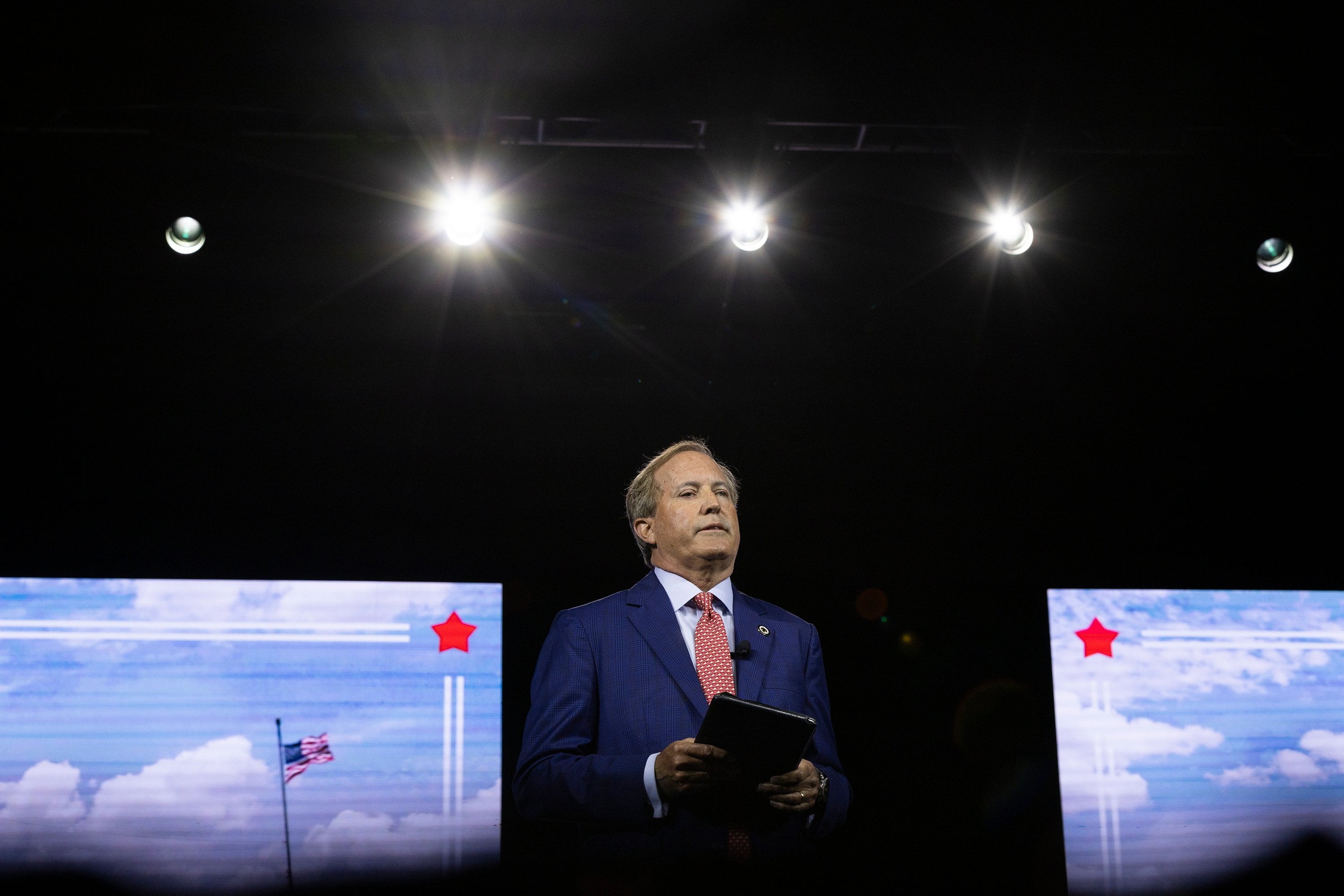 A man in a blue suit stands on a darkened stage between two screens, each showing an American flag